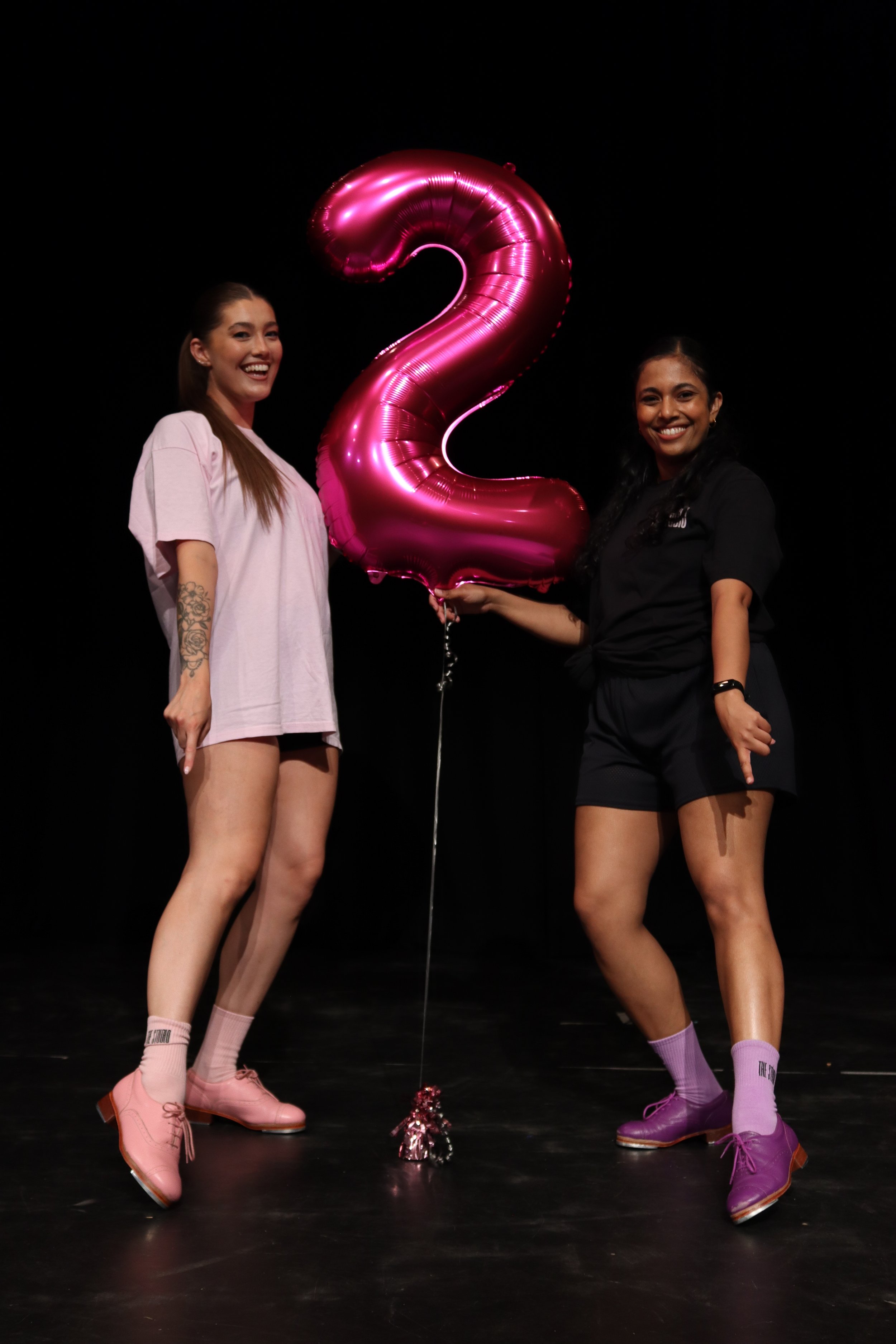 Two smiling women standing on a stage, holding a large pink foil balloon shaped like the number two, with a black background. They are wearing casual clothing and colorful socks and shoes.