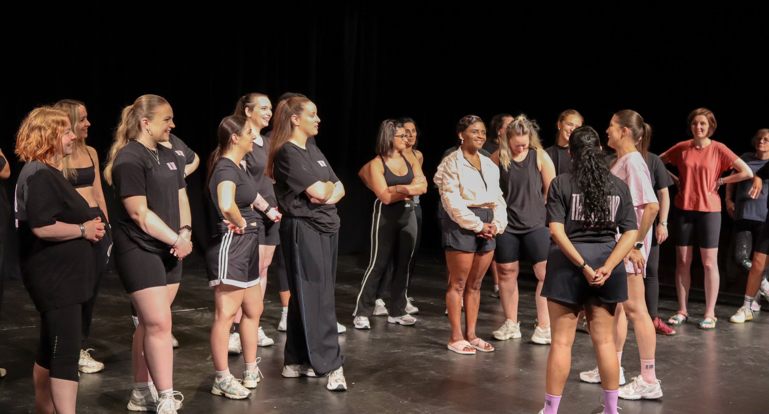 A group of women on stage, standing in a semi-circle, engaged in conversation or rehearsal, dressed in casual and workout attire, with a black background.