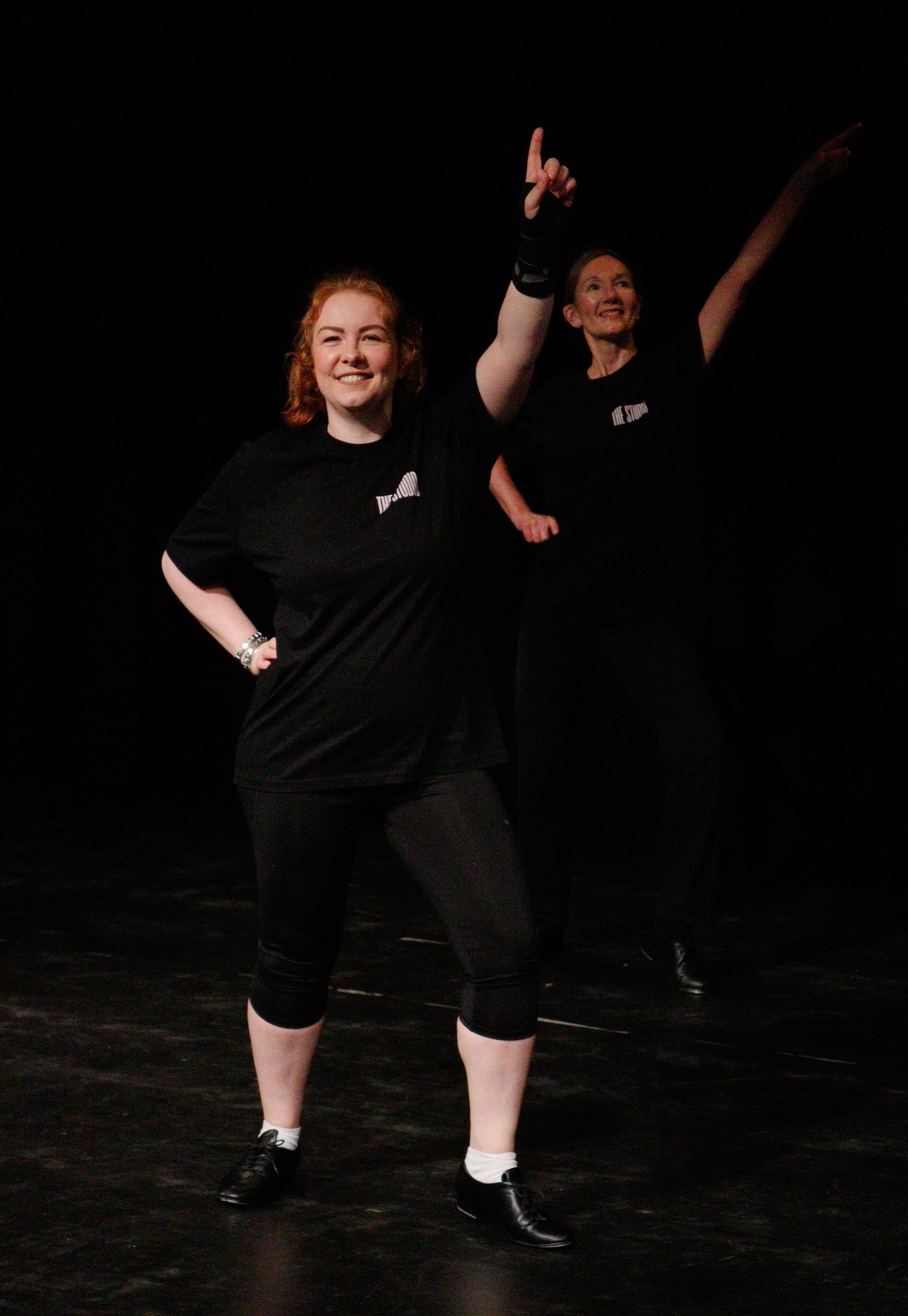 Two women in black t-shirts and black pants with feet apart dancing on a dark stage, smiling and pointing upward with their arms raised.