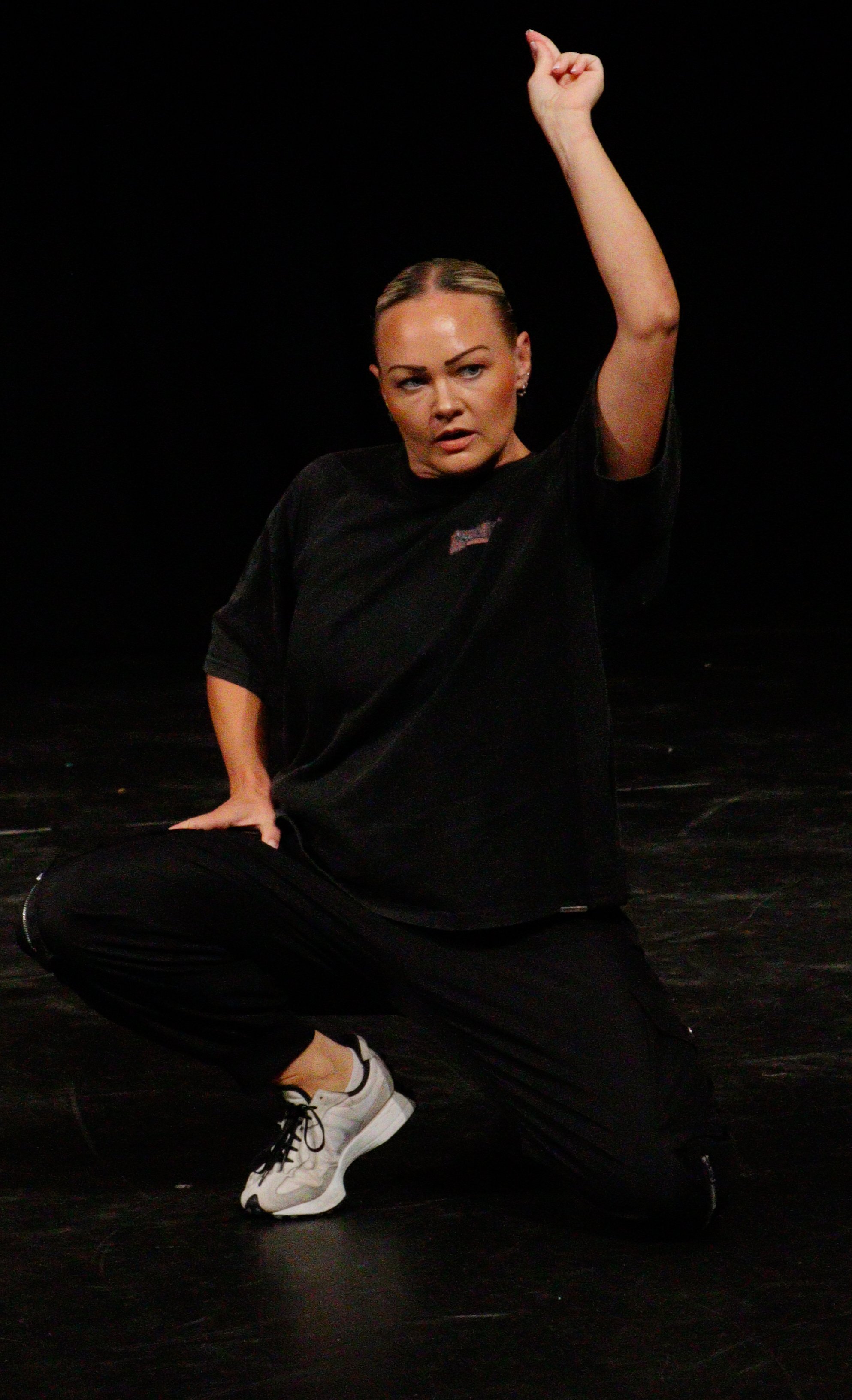 A woman with blonde hair in a bun, wearing a black T-shirt and black pants, is sitting on the floor with one knee up. She is raising her right arm with a clenched fist and has a focused, intense expression.