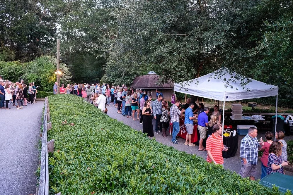 People standing in line outdoors beside a food stand under a white canopy, with a backdrop of trees and a small wooden shelter.