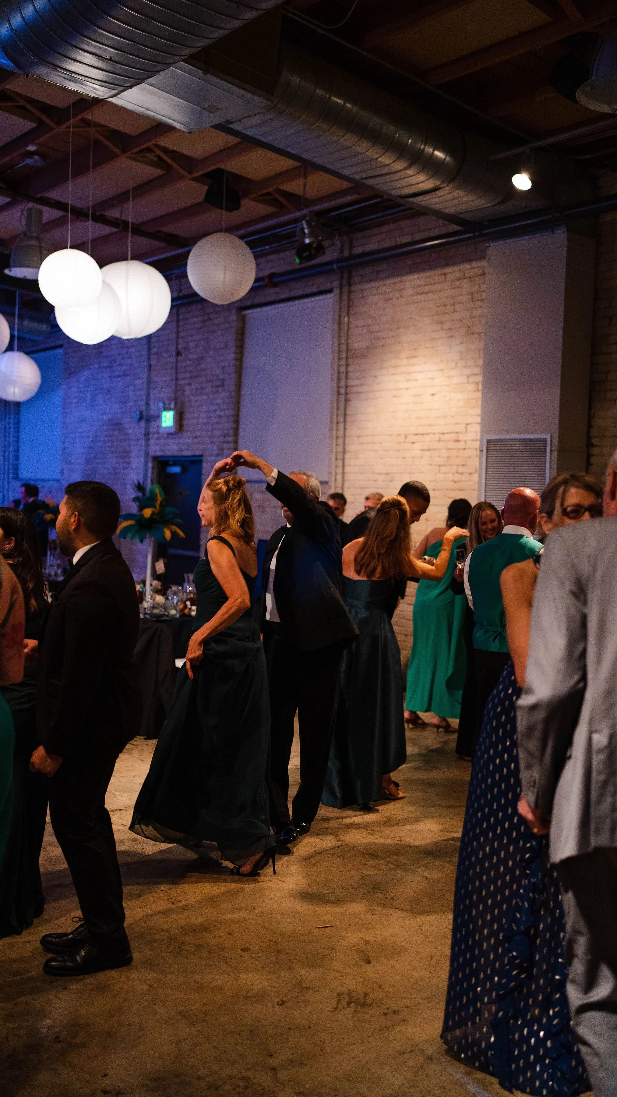 People dancing and socializing at an indoor event with dim lighting, lantern ceiling lights, brick walls, and a concrete floor.