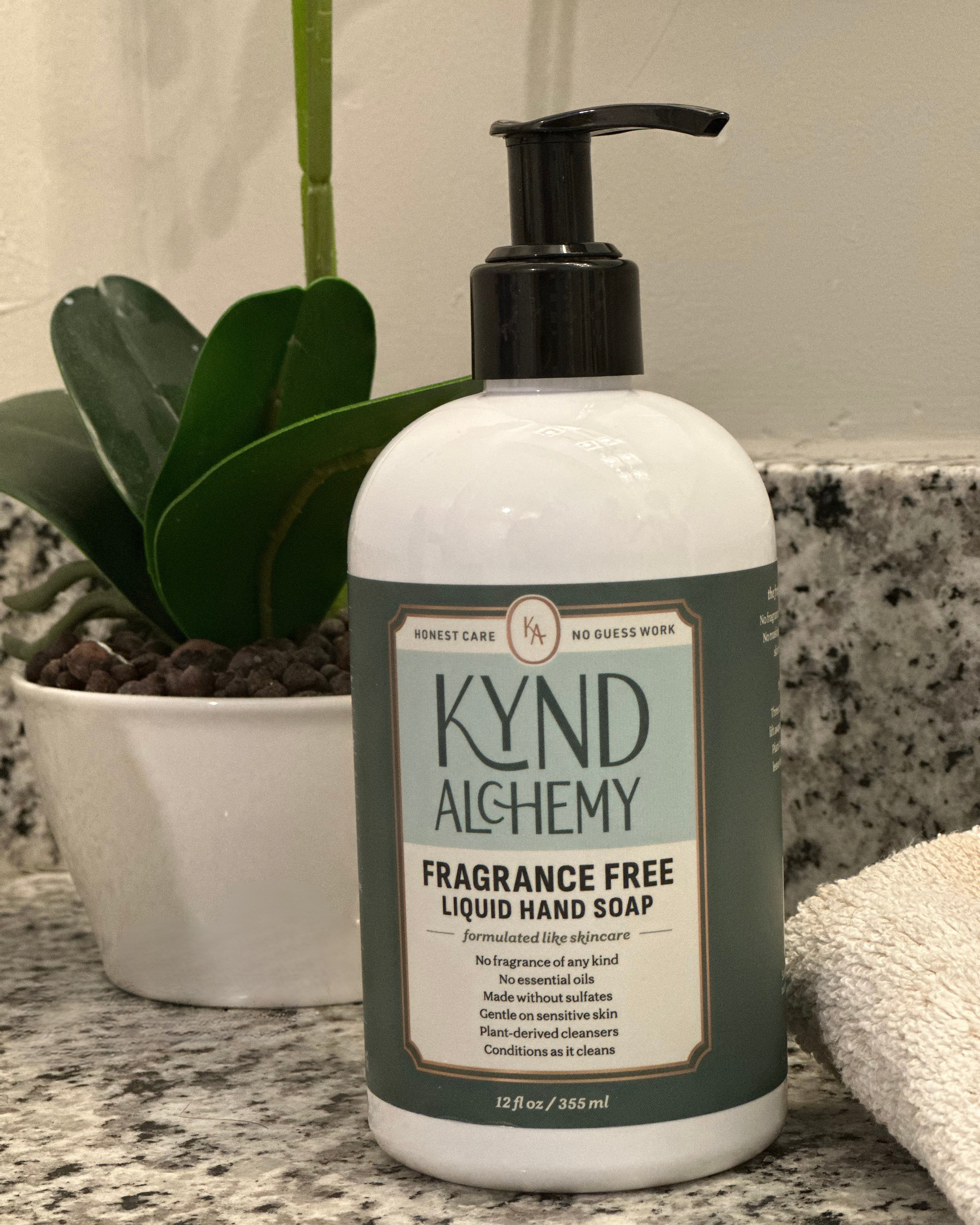 Kynd Alchemy 12 oz bottle seating on a bathroom counter next to a plant and a hand towel