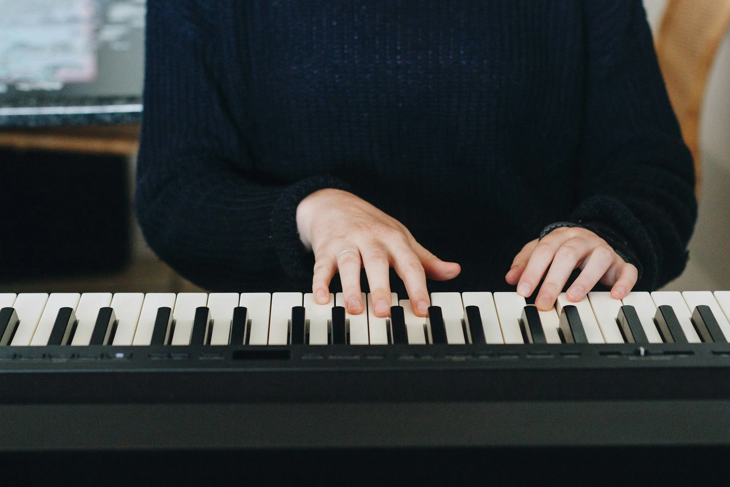 Person playing a keyboard, wearing a black sweater, with hands on the keys.