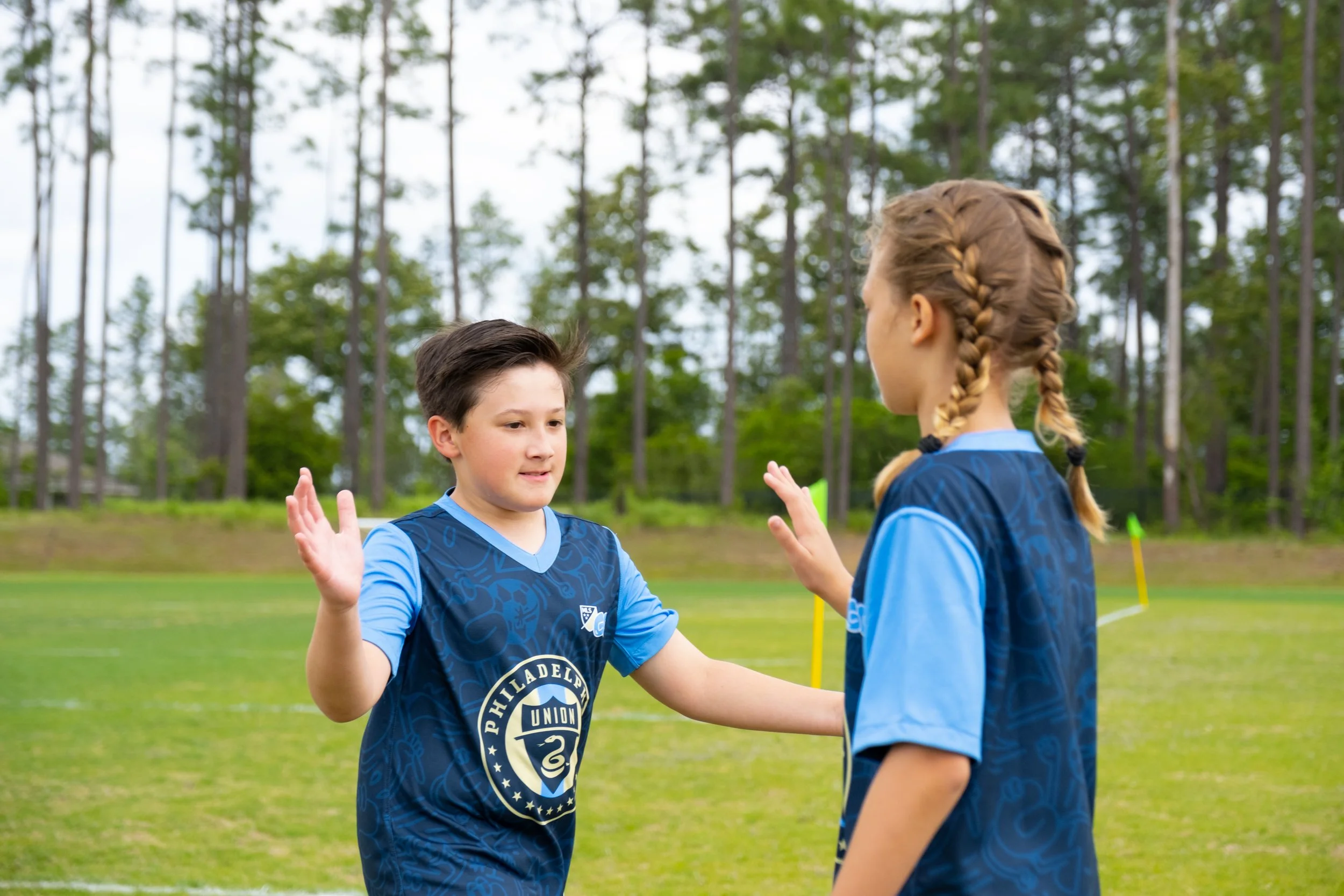Two young boys wearing blue soccer uniforms greeting each other with high fives on a soccer field surrounded by trees.