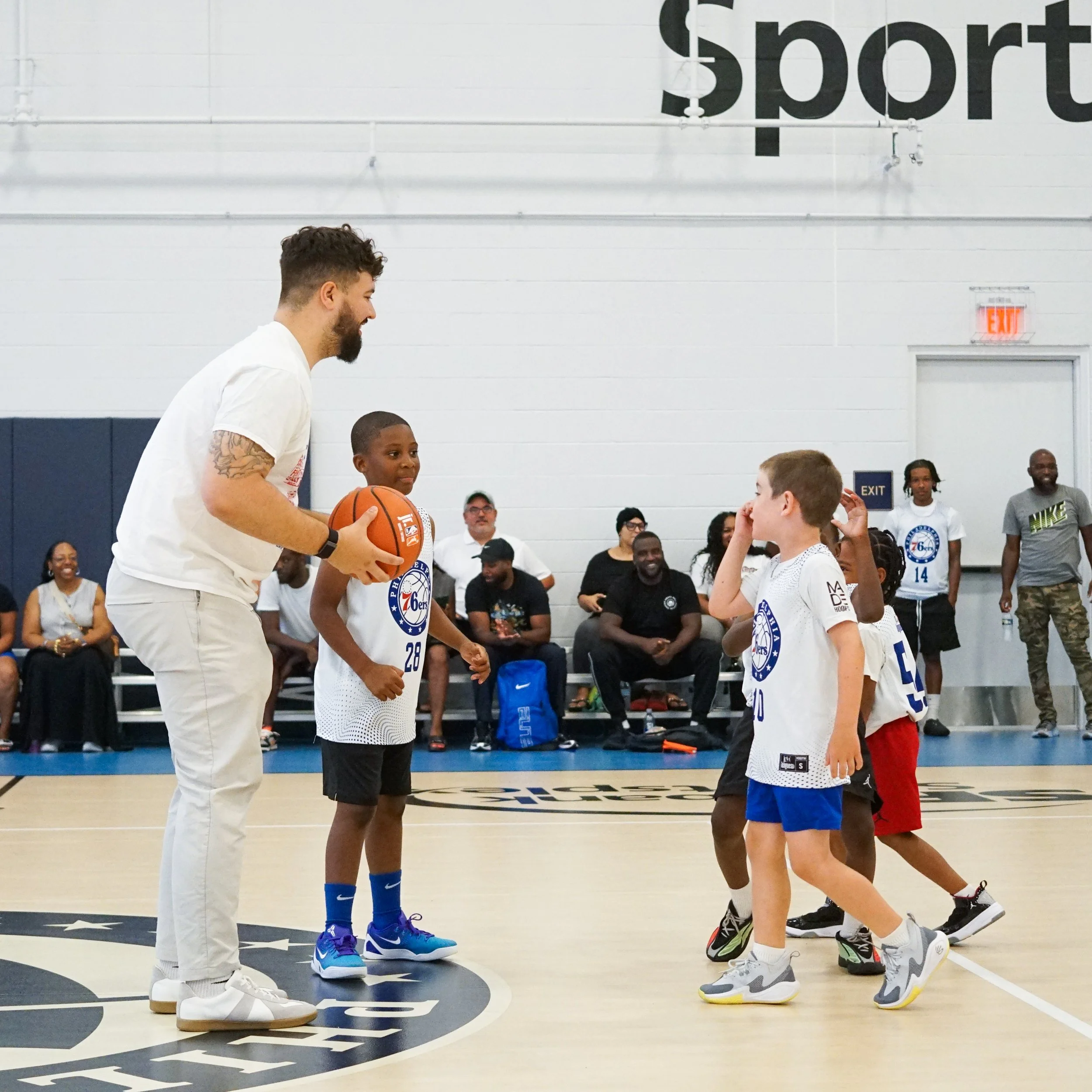A man with a beard and tattoos on his arm is coaching young boys during a basketball practice or game inside a gymnasium. The man is holding a basketball and appears to be instructing the children, who are focused on him. Several adults and children are watching in the background.