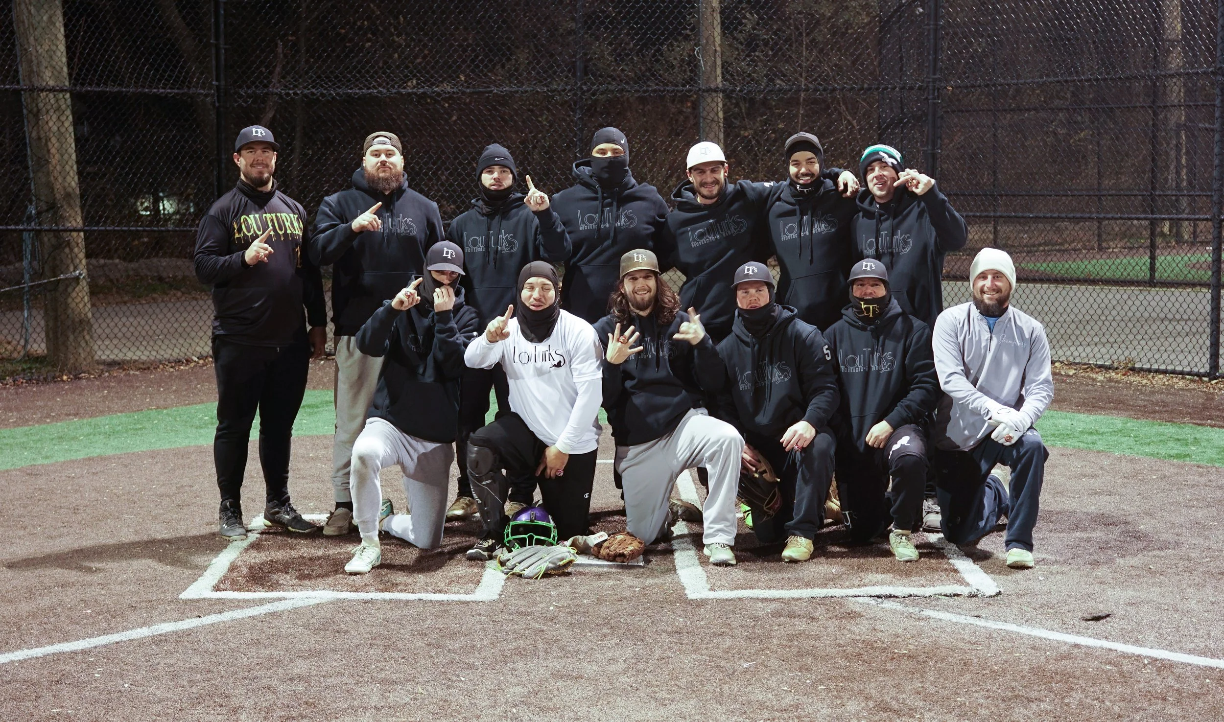 A group of twelve men posing together on a baseball field at night. They are wearing black or gray hoodies, jackets, and baseball caps. Some men are making hand gestures, and one man is in front holding a baseball glove and a baseball. Behind them is a chain-link fence and a dark forested background.