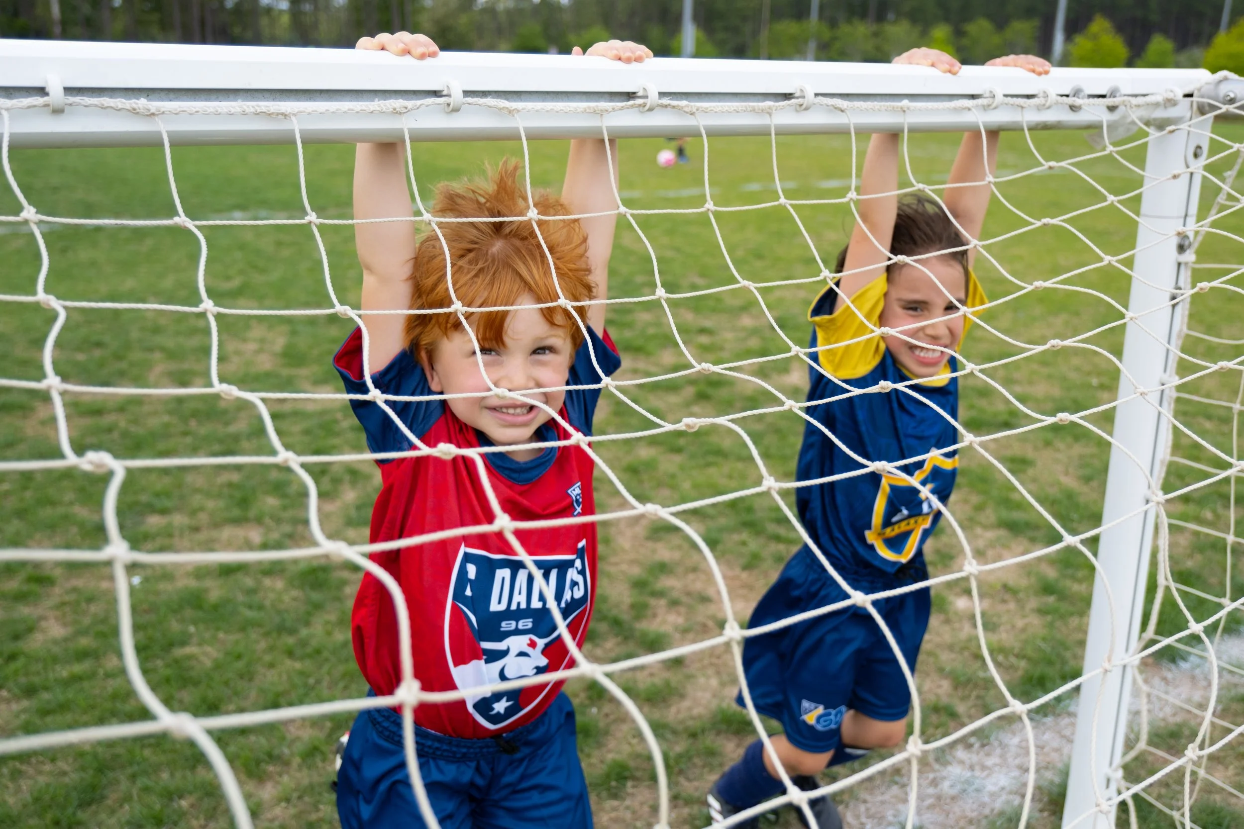 Two children playing soccer, hanging upside down on a soccer goal net in a grassy outdoor field.