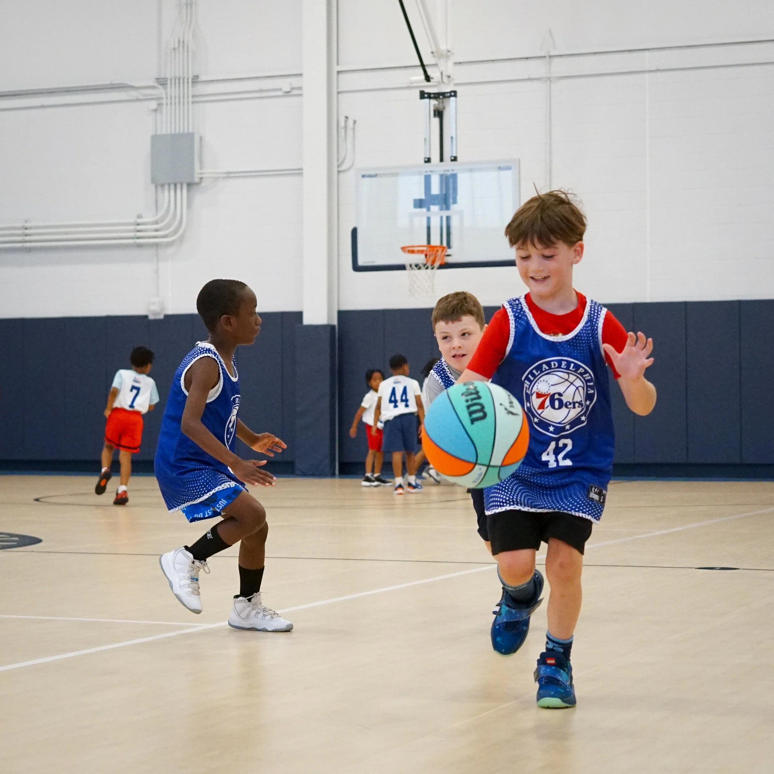 Children playing basketball in an indoor gym, wearing sports uniforms.