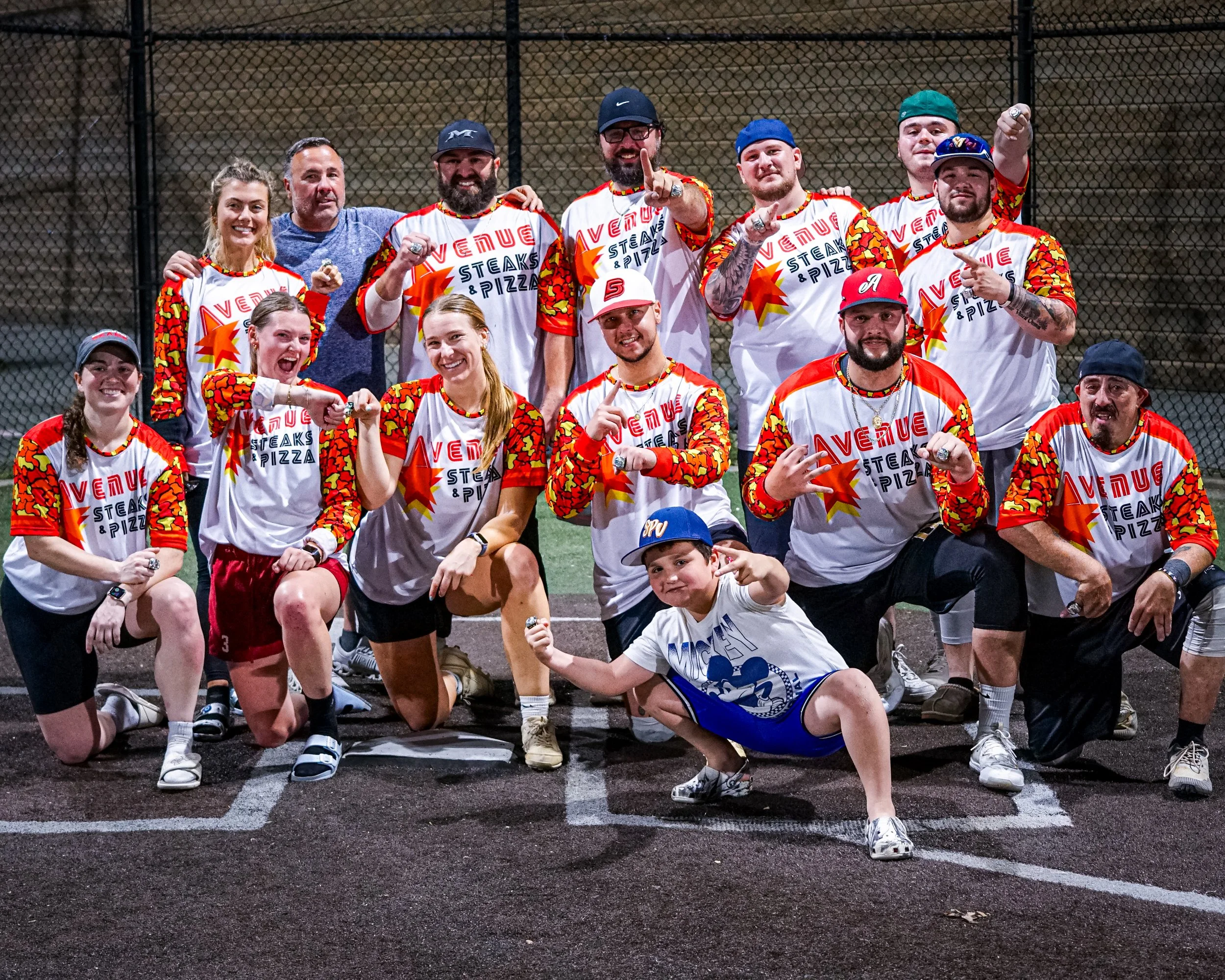 Group of people posing on a baseball field at night. They are wearing matching sports jerseys with 'Venture Steaks & Pizza' logo, some raising fists and pointing. They are smiling and kneeling or standing in front of a chain-link fence.