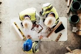 Three construction workers wearing helmets and safety gear working at a table with tools and blueprints.