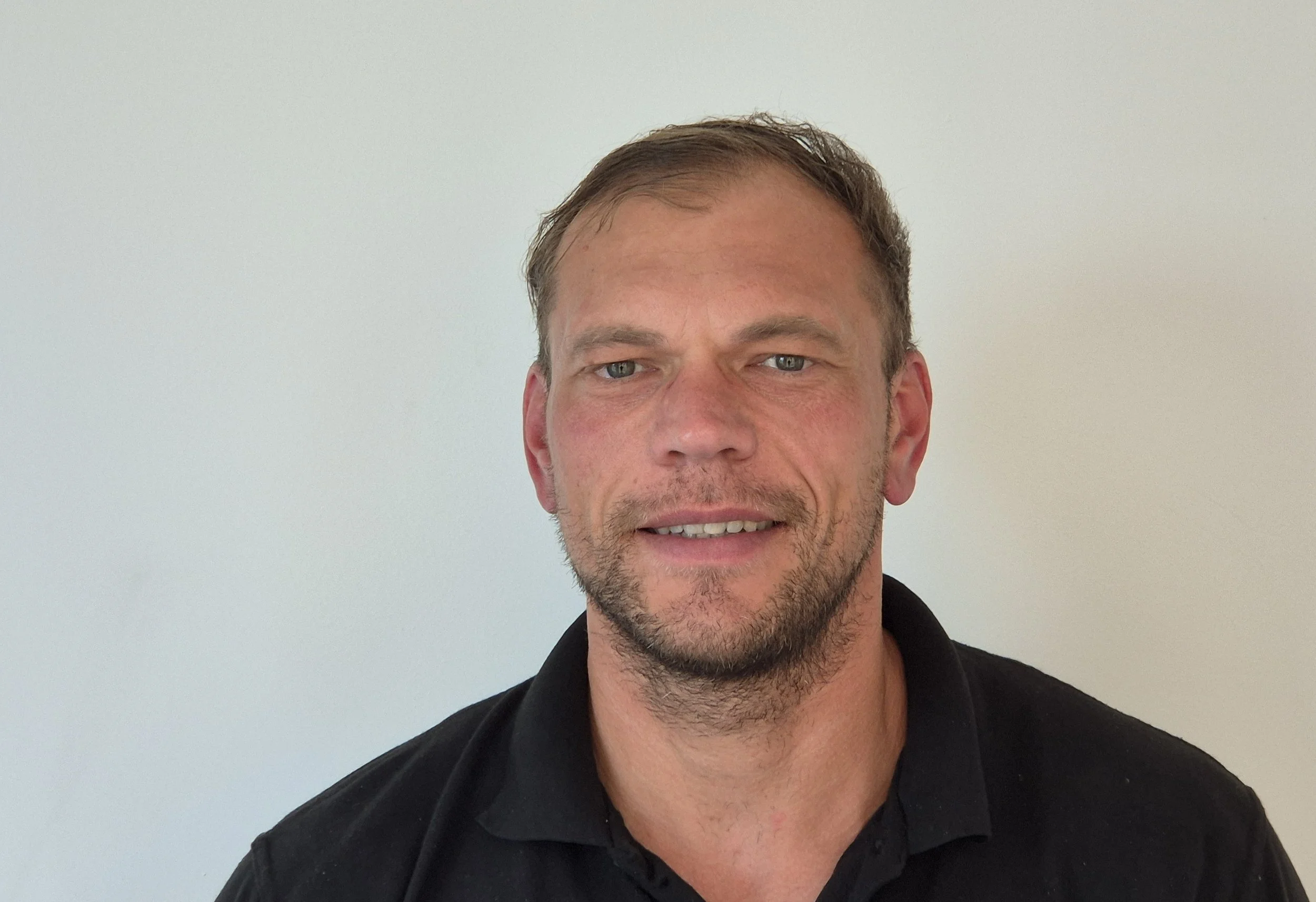 A man with light skin, short brown hair, and a beard, wearing a black shirt, standing against a plain white wall.