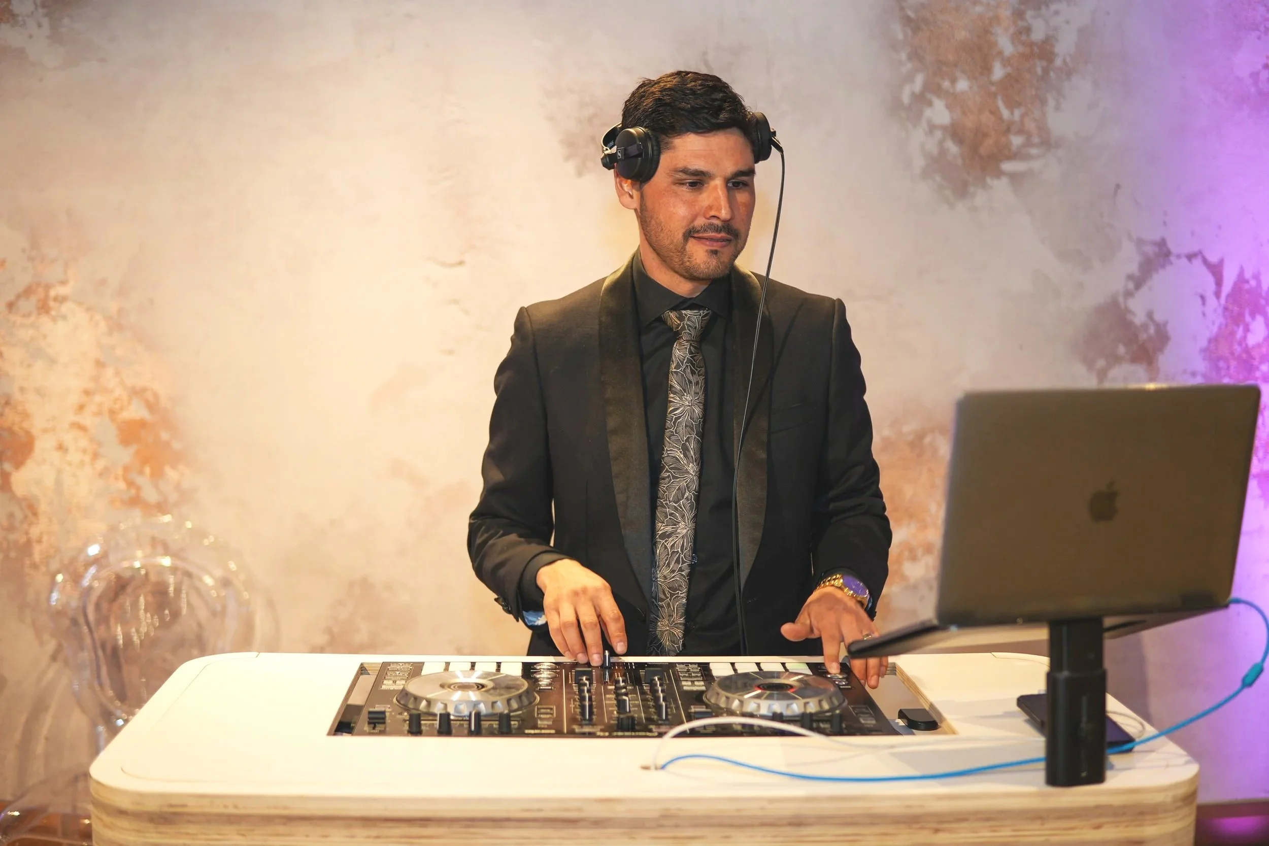 A man in a black suit and patterned tie DJing at a white table with a DJ controller, wearing headphones, in a setting with a colorful textured wall.
