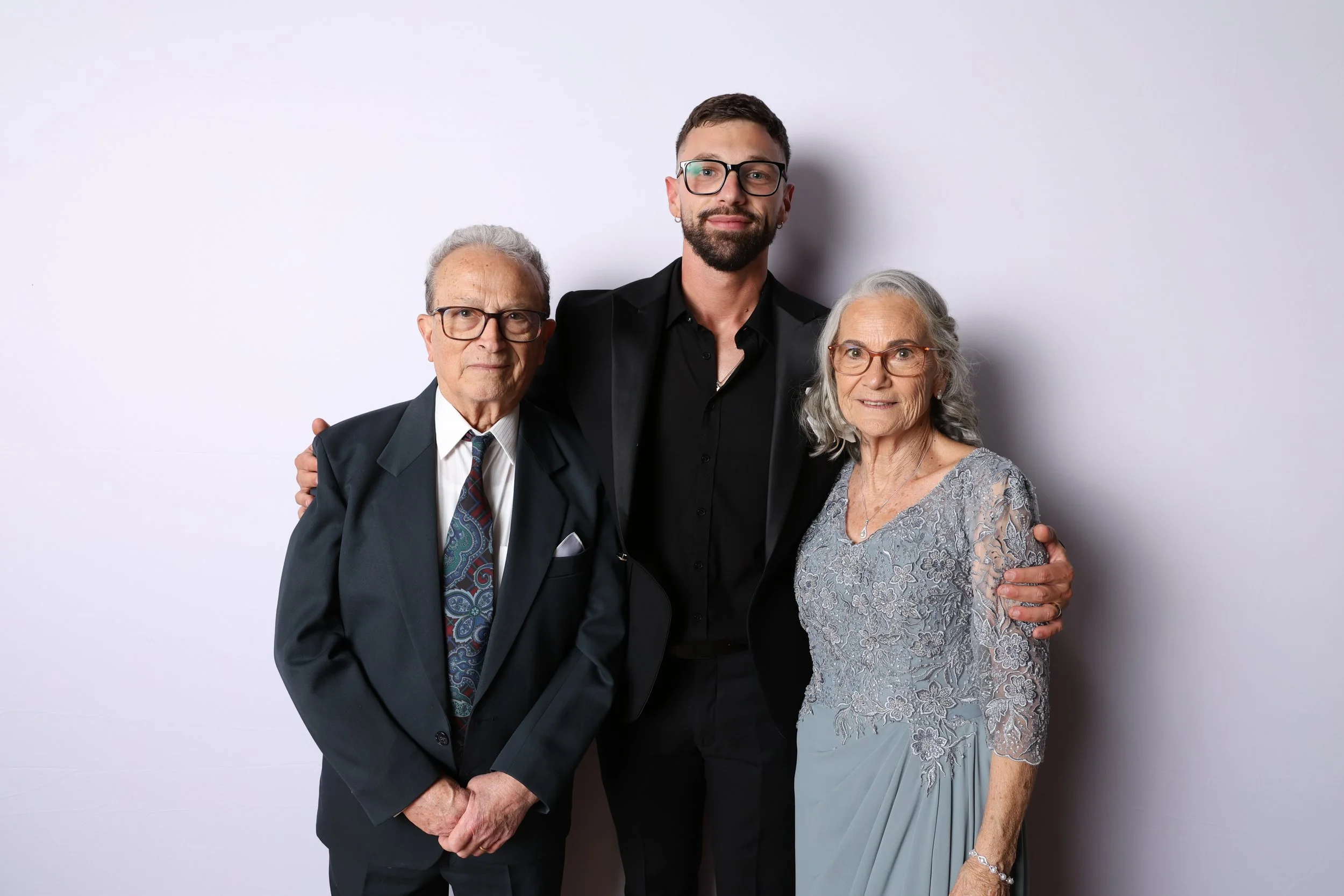 A young man with glasses and dark hair standing between an elderly man and woman, all smiling and dressed formally, against a plain light background.