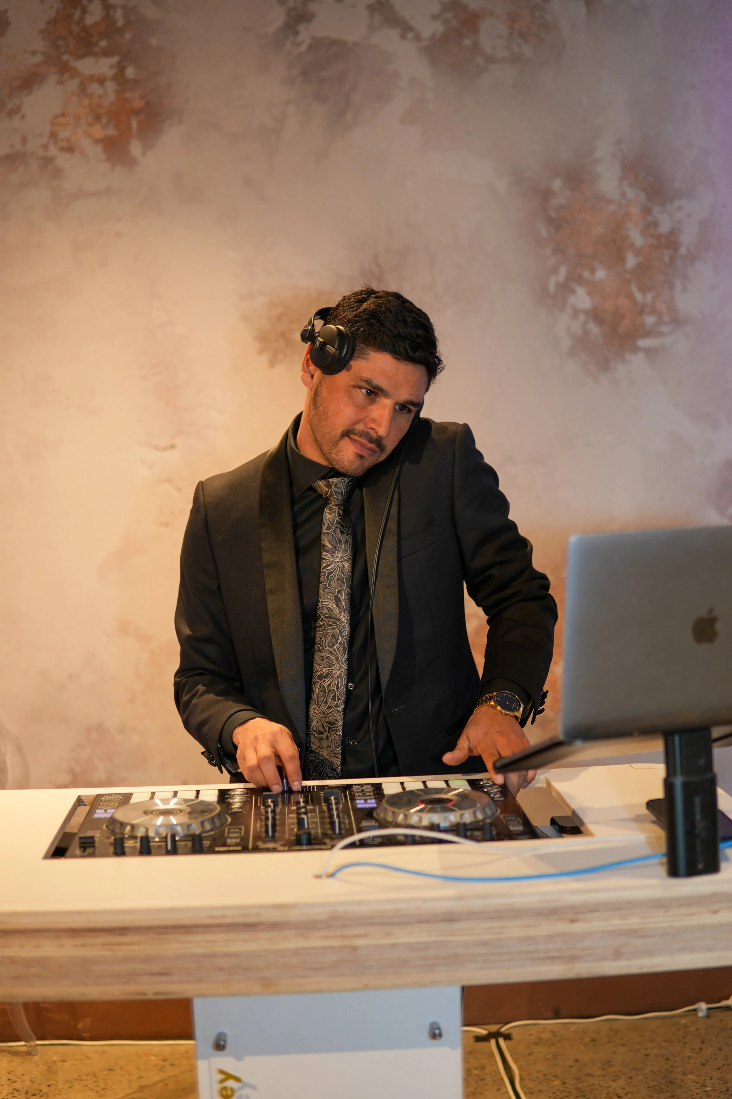 A man in a black suit with a patterned tie and gold watch DJing with turntables and a laptop, wearing headphones, at a white table against a textured beige and purple wall.