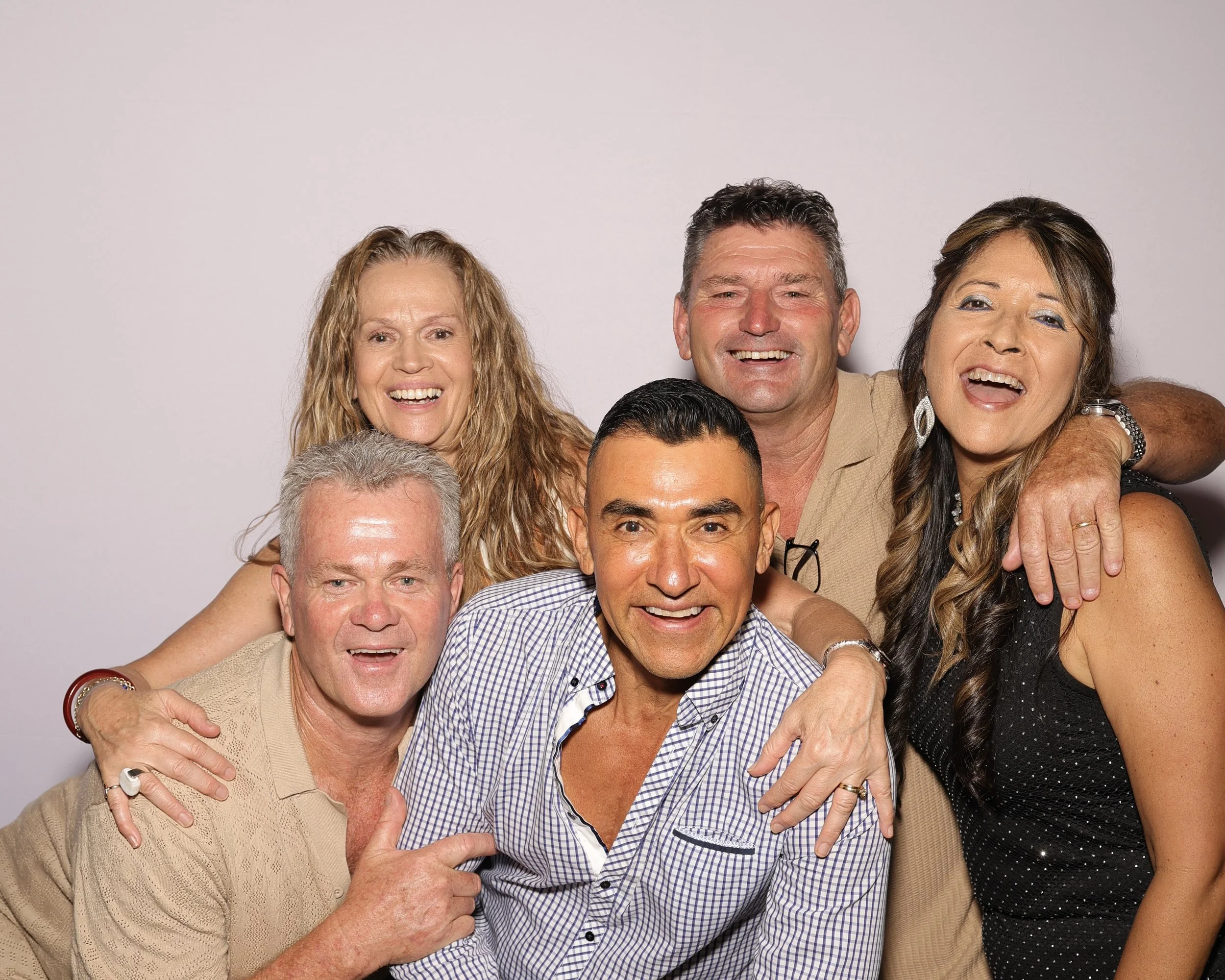 Group of smiling, diverse adults close together, embracing and looking at the camera against a plain background.