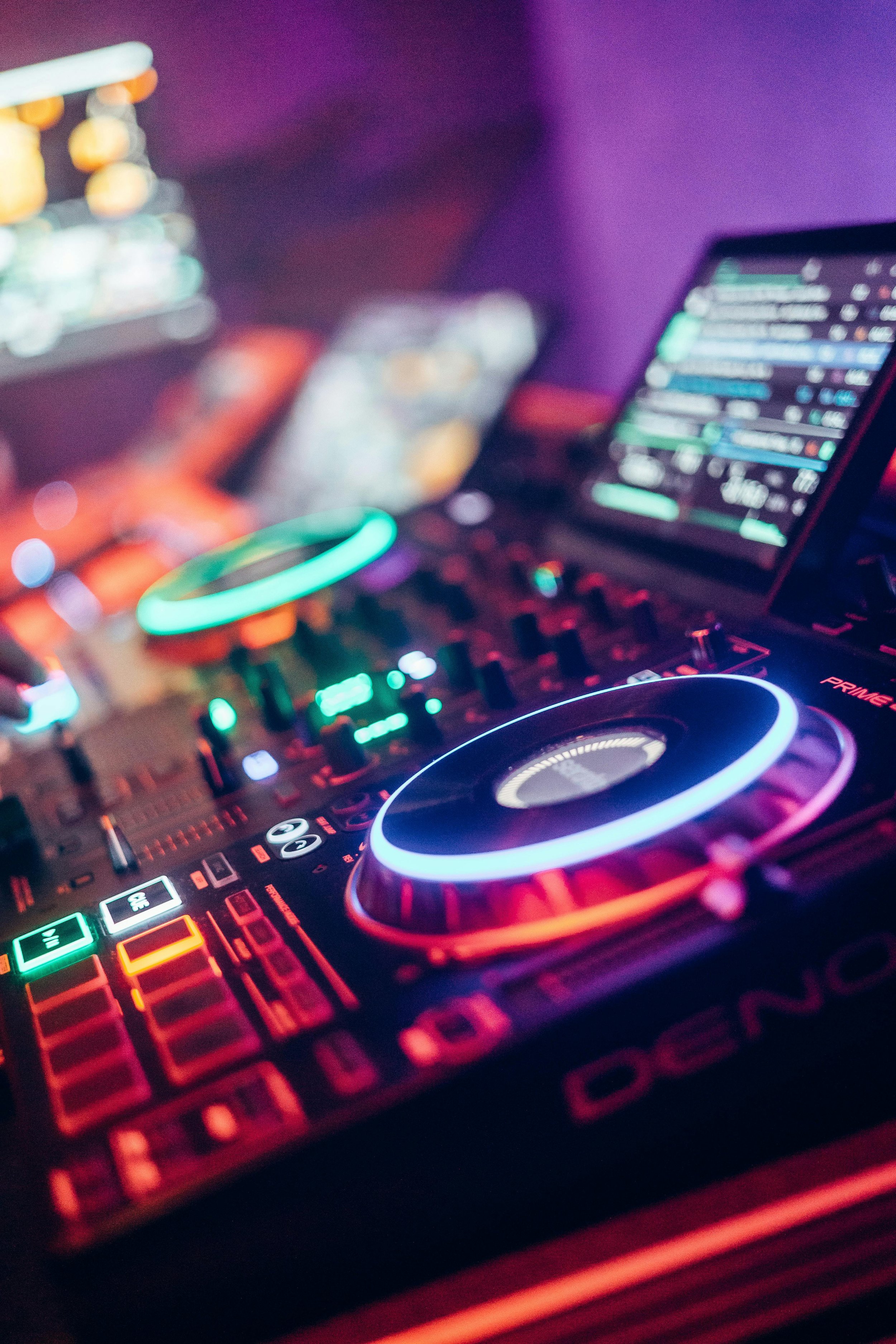 Blurred photo of a DJ mixing music with colorful LED lights, a laptop, and a MIDI controller in a dark, or nightclub setting.