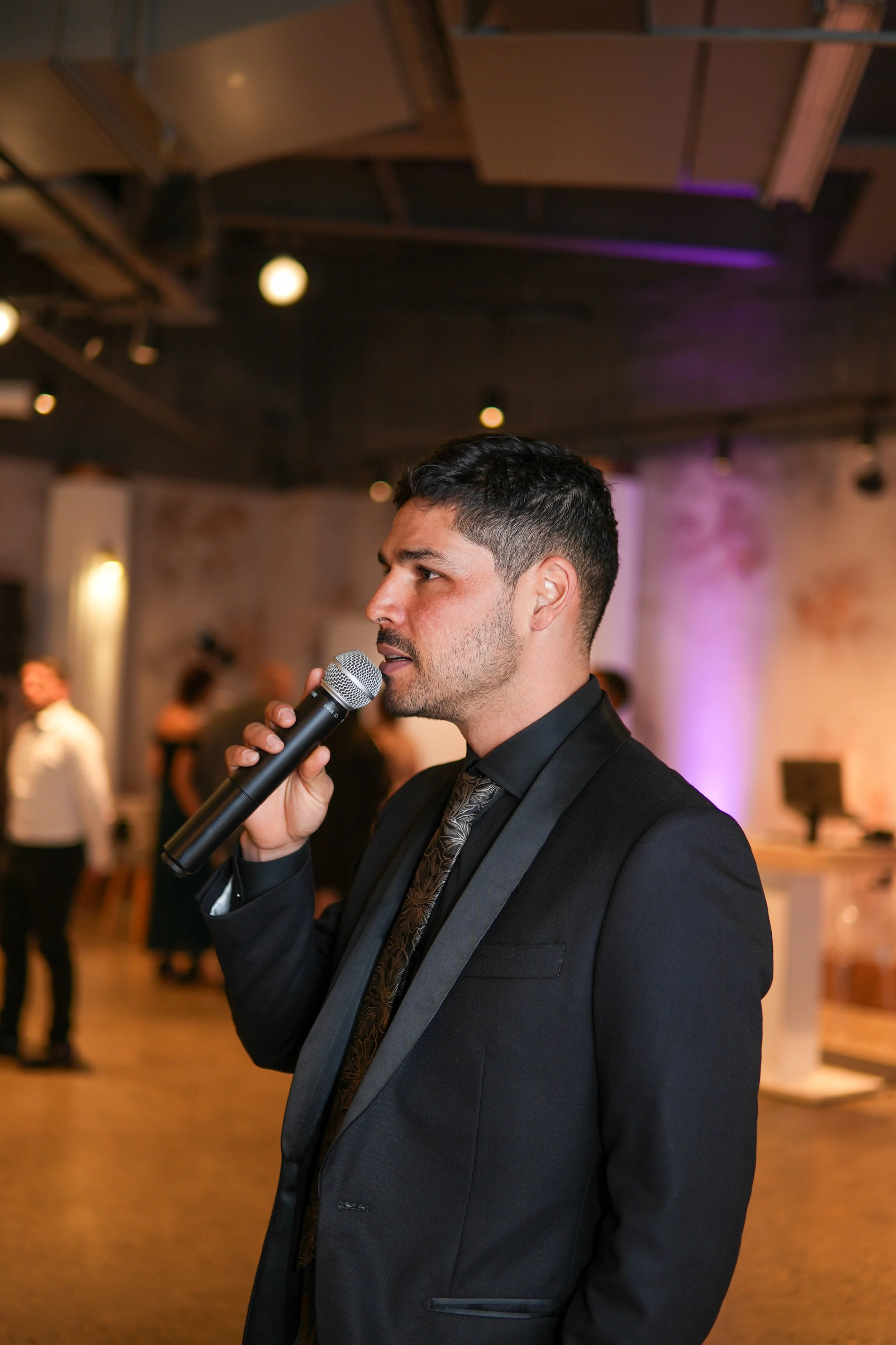 A man in a black suit and tie speaking into a microphone at an indoor event with a brick wall and blurry people in the background.