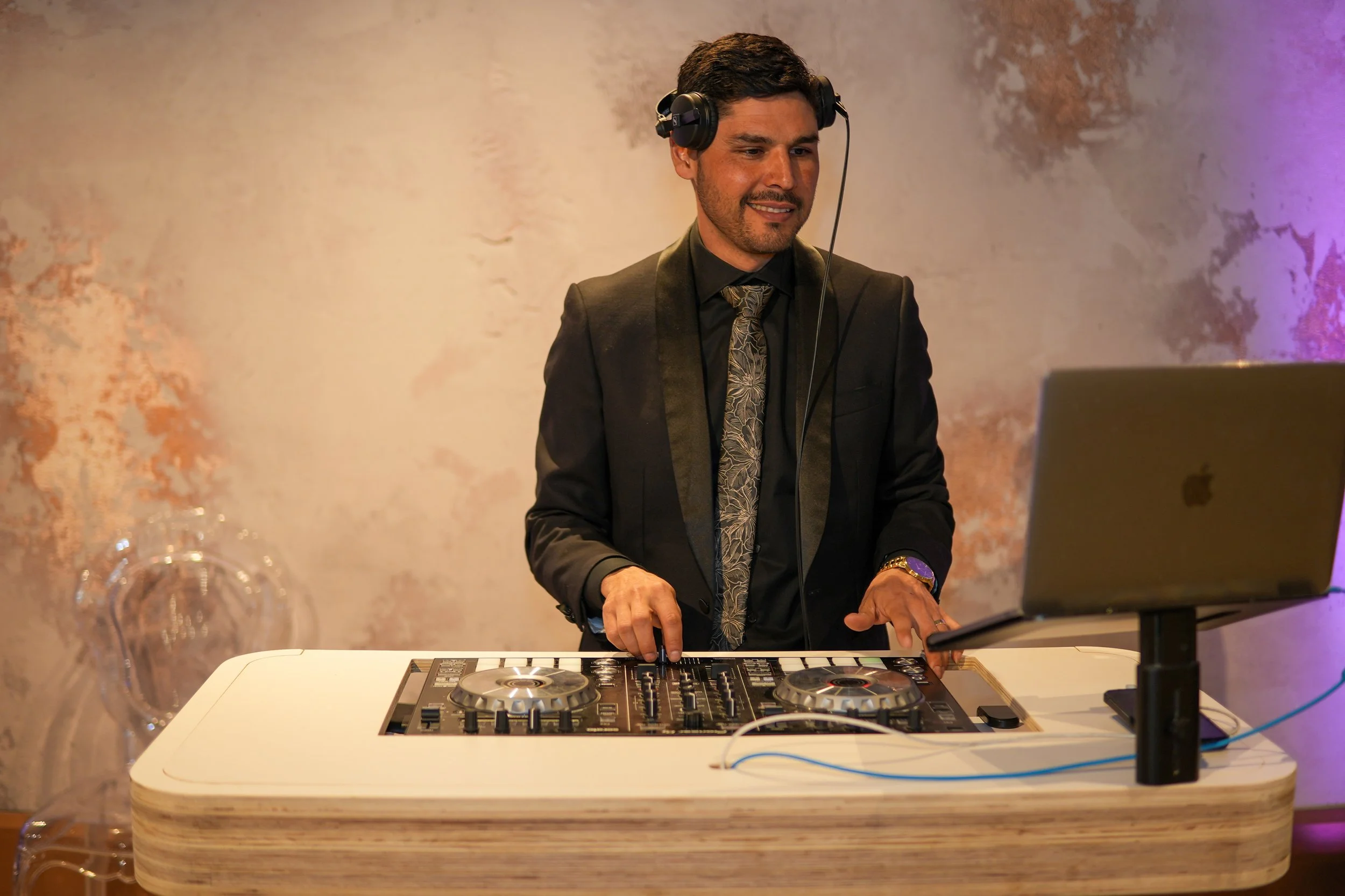 A man in a black suit and patterned tie mixing music on a DJ controller at an event, with a background featuring a textured wall with pink and purple lighting.