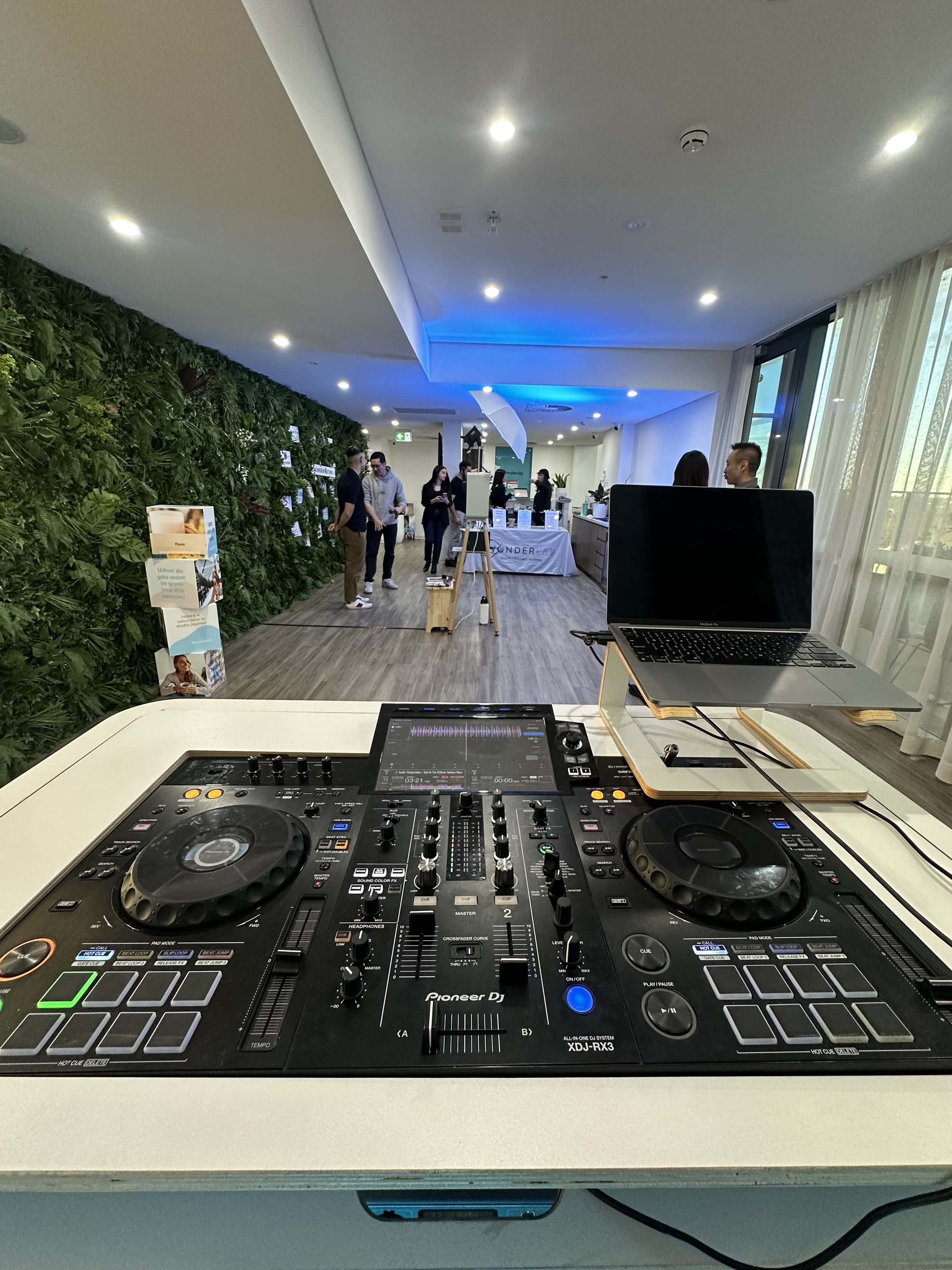 DJ turntable and mixer setup on a table in a room with people mingling, green wall on the left, large windows on the right, and a reception or display area in the background.