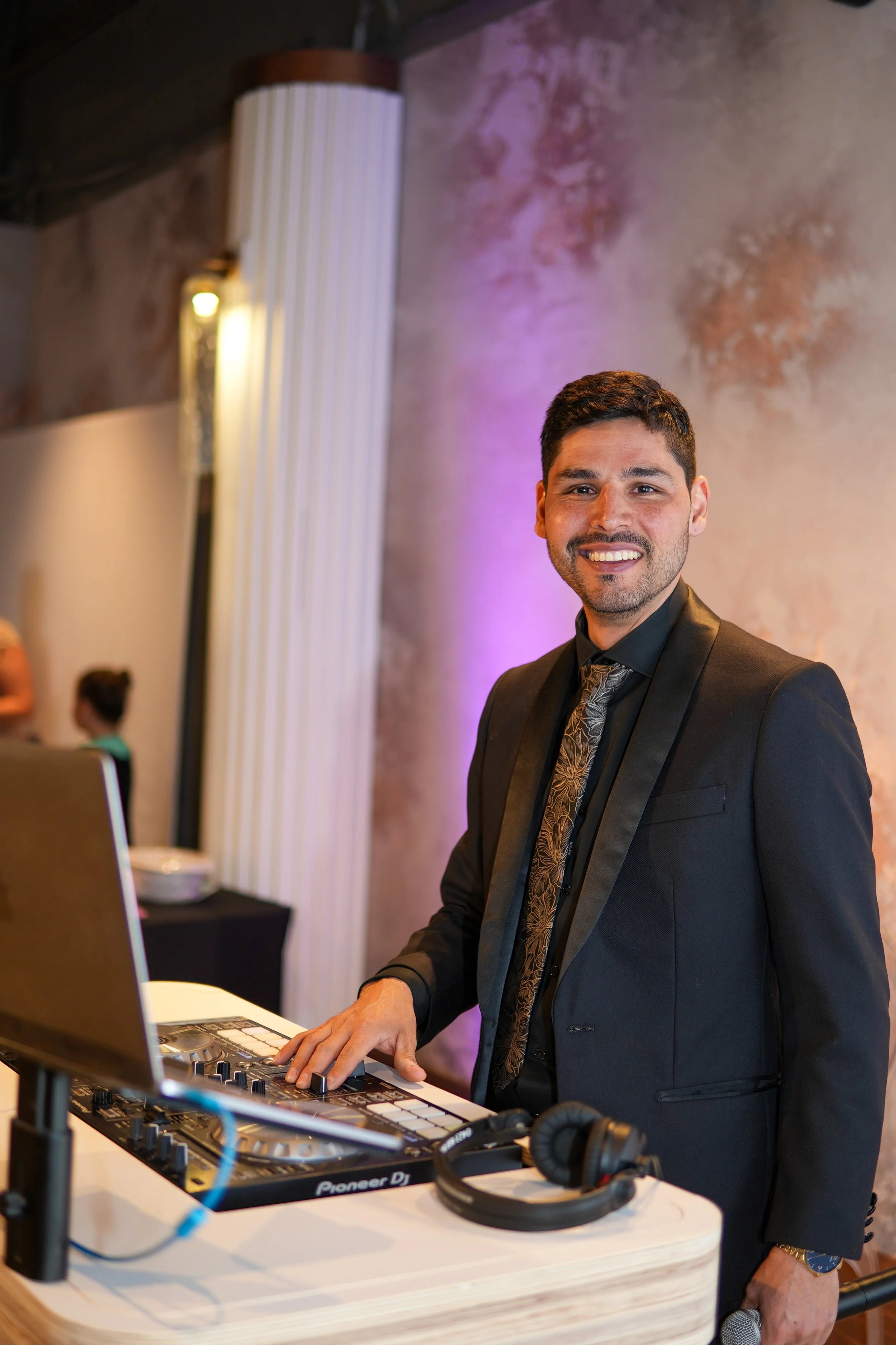 A man in a black suit and tie is DJing at an event, smiling, with a DJ controller and headphones in front of him. The background features a textured wall with purple lighting.