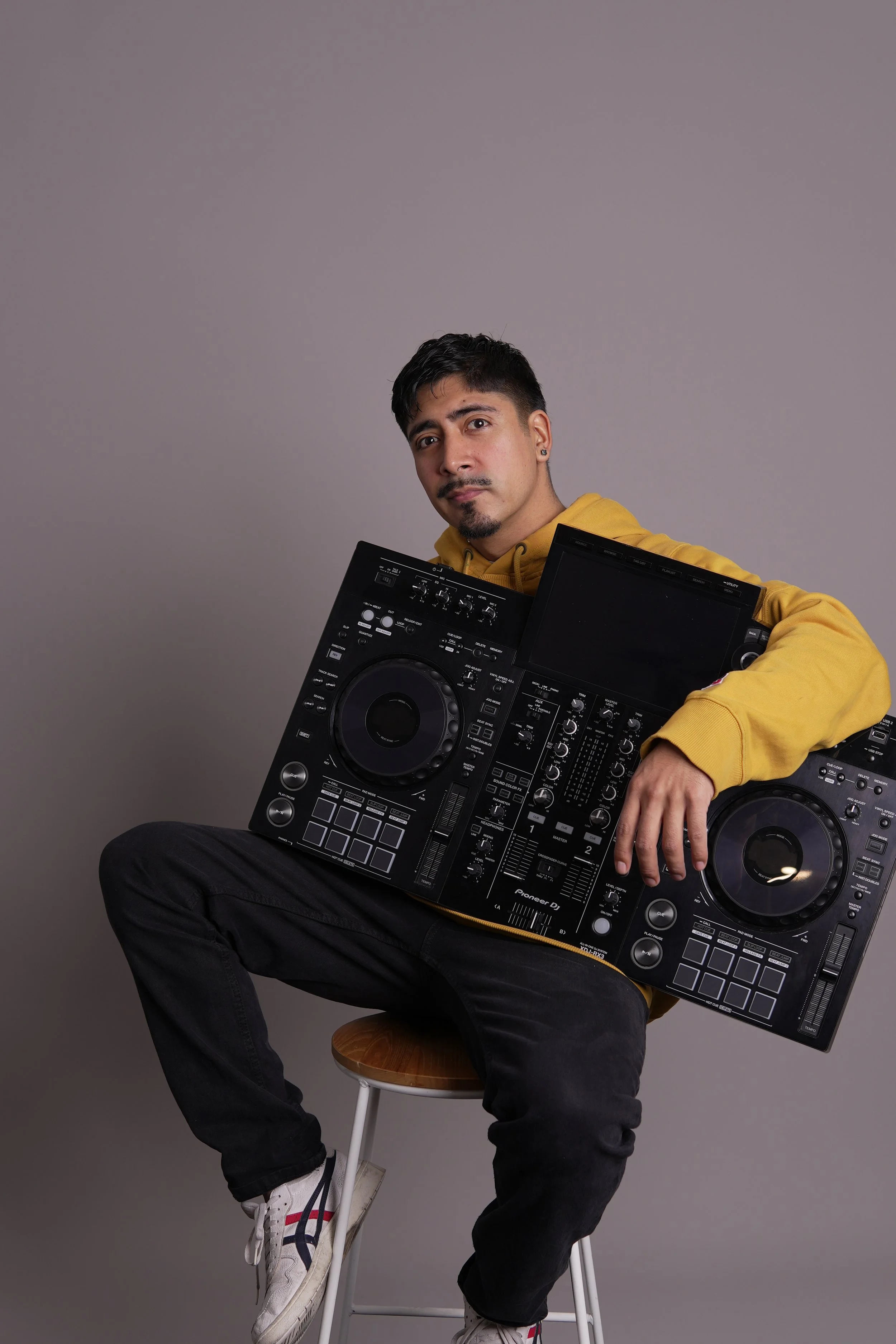 A man sitting on a stool, holding a DJ controller, wearing a yellow hoodie and black pants, against a gray background.