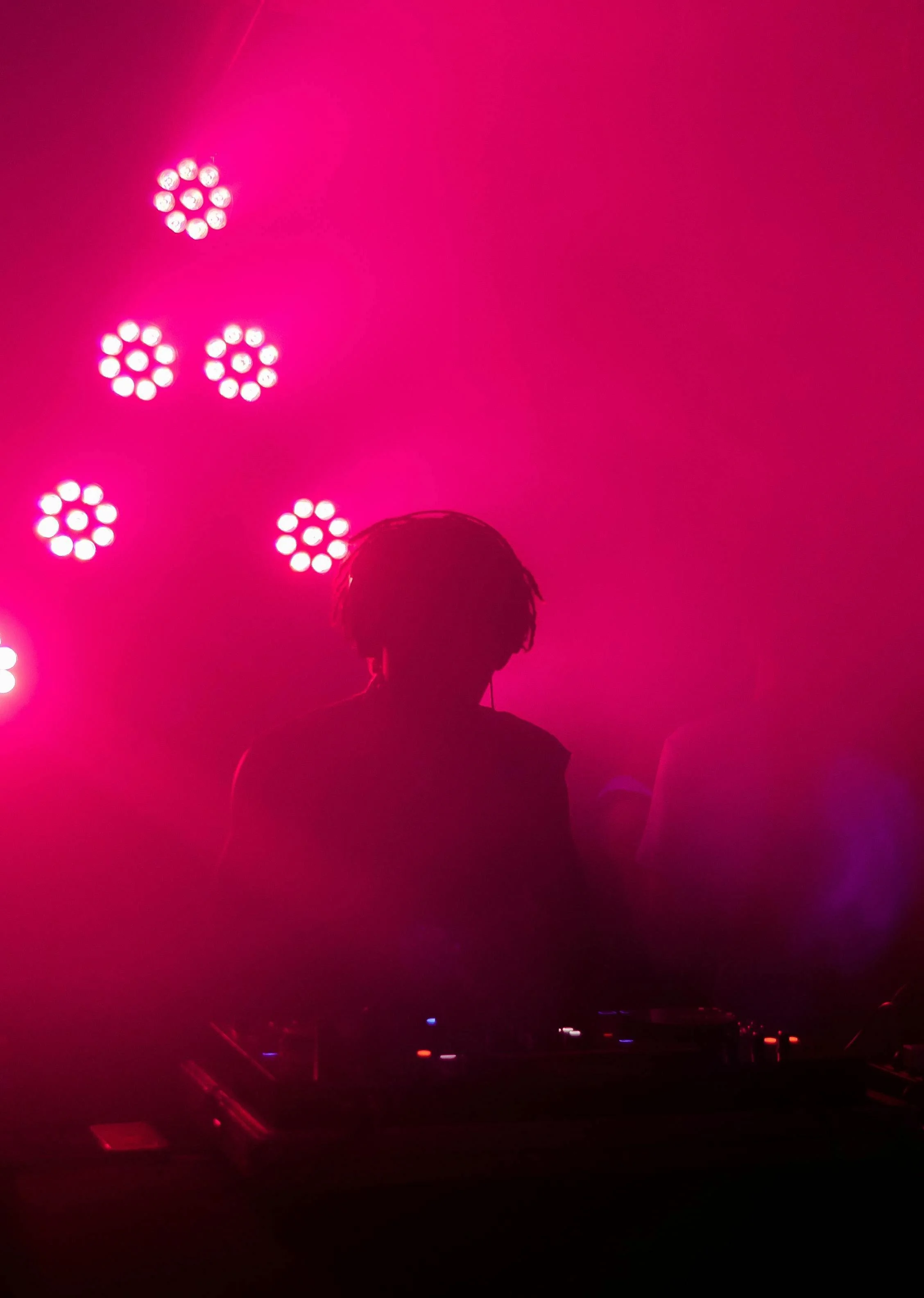 Silhouette of a DJ with pink lighting and circular LED stage lights in the background, mixing music at a nightclub or concert.