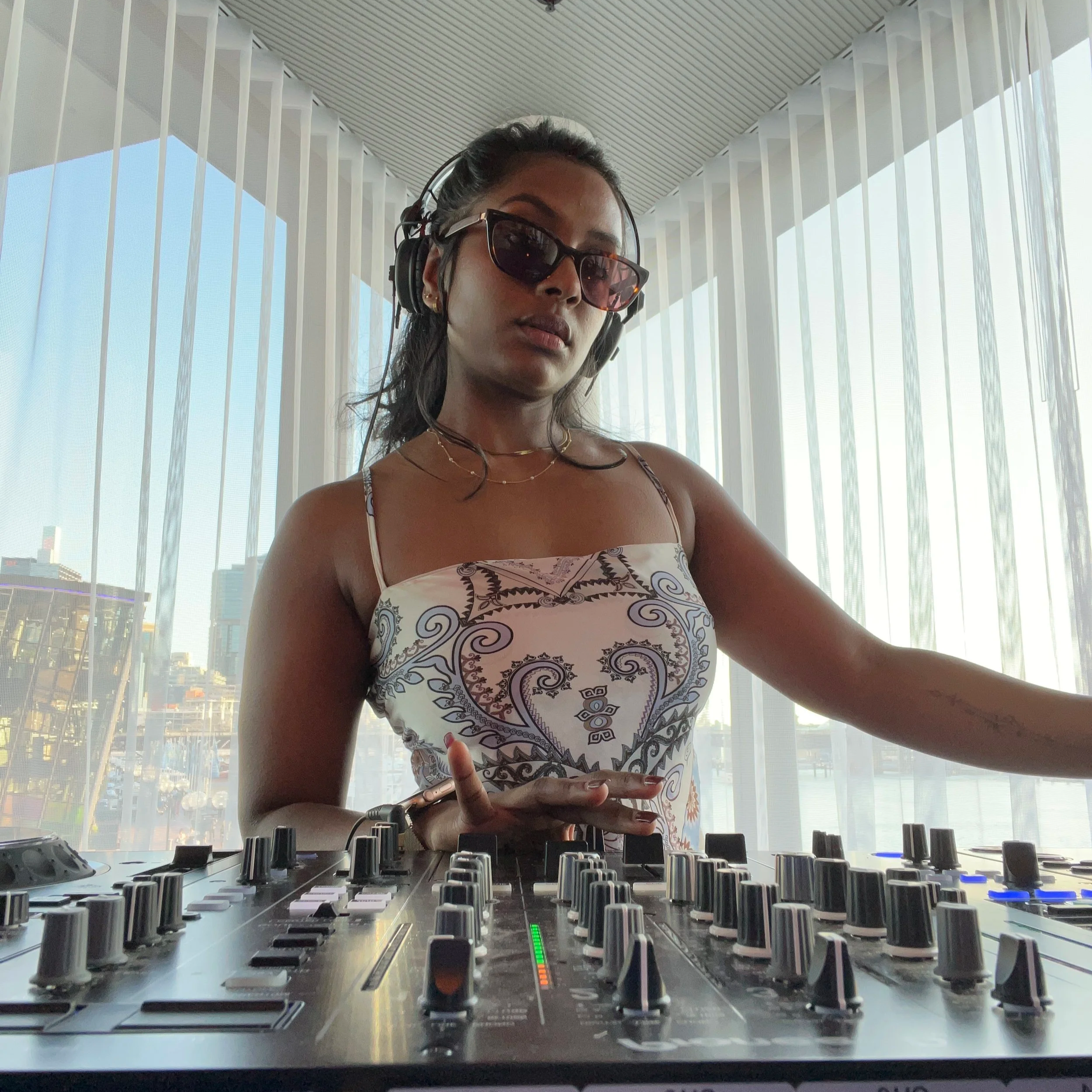 A woman wearing sunglasses, headphones, and a patterned dress, standing in front of a DJ mixer with city buildings and blue sky visible through large windows behind her.