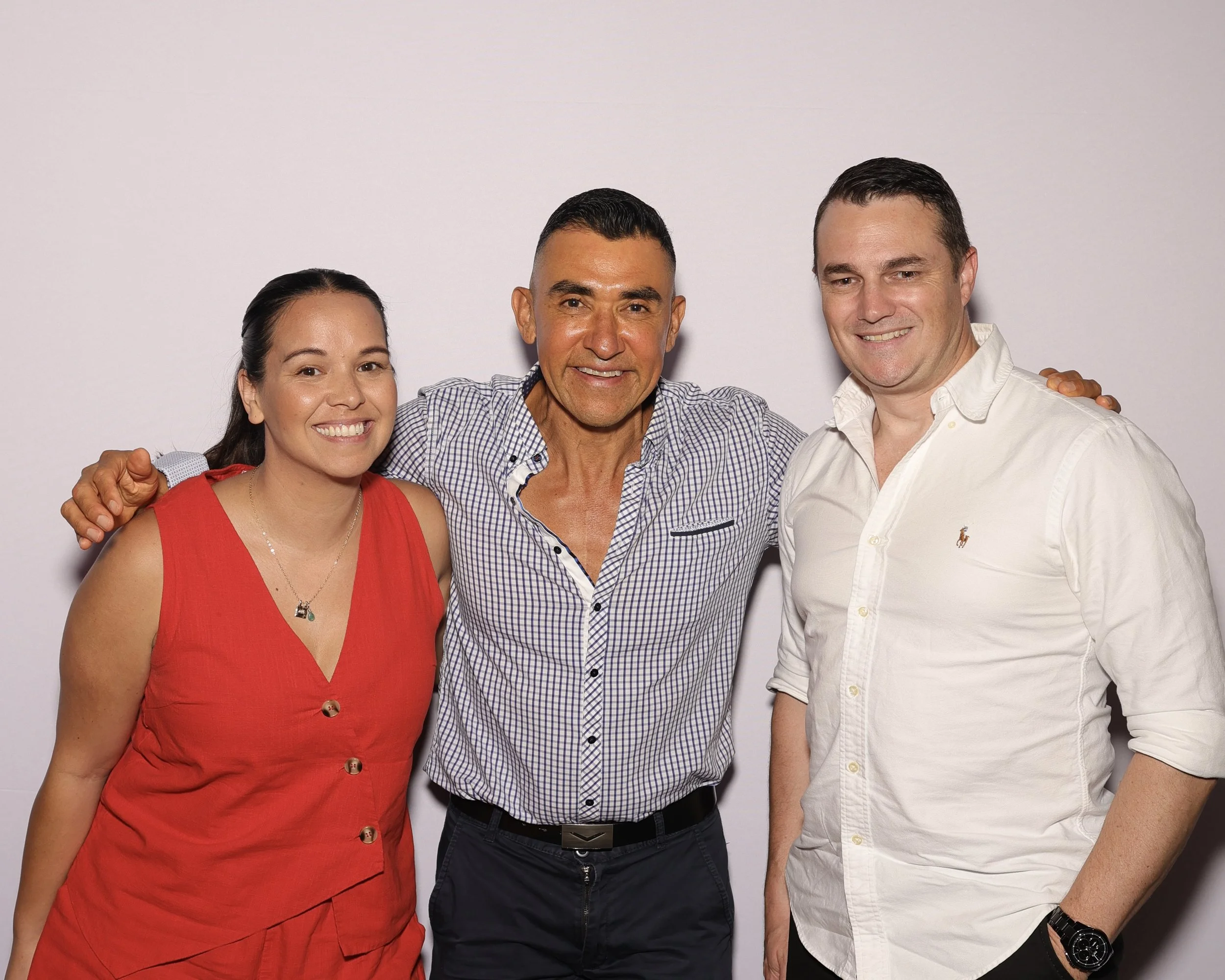 Three people smiling and posing together against a plain white background. The woman on the left wears a red sleeveless dress, the man in the middle wears a checked shirt, and the man on the right wears a white shirt with rolled-up sleeves.