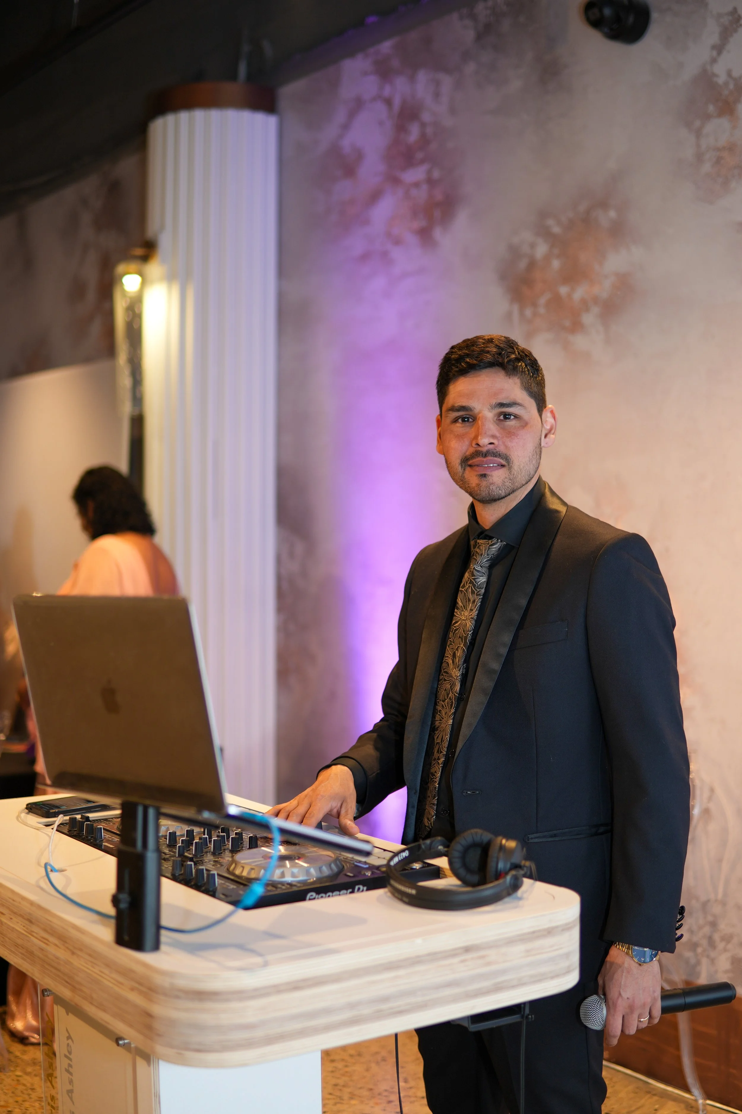 A man in a black suit and patterned tie is DJing at an event, standing behind a DJ console with a laptop and headphones on a wooden table, holding a microphone in his right hand, with a textured wall and a woman with curly hair and a peach-colored dr
