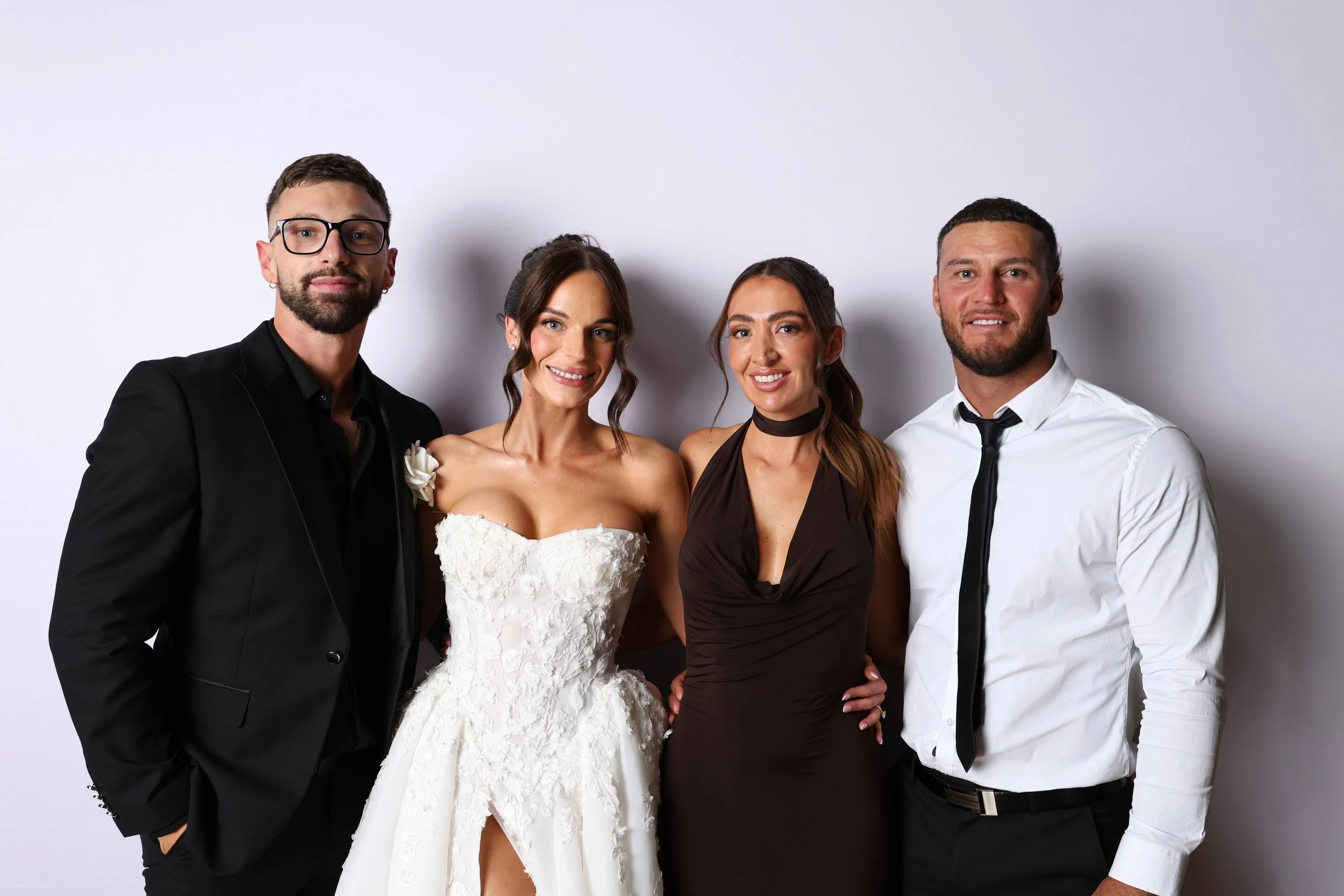 Four people posing together against a plain white background, including a woman in a wedding dress and three men in formal attire.