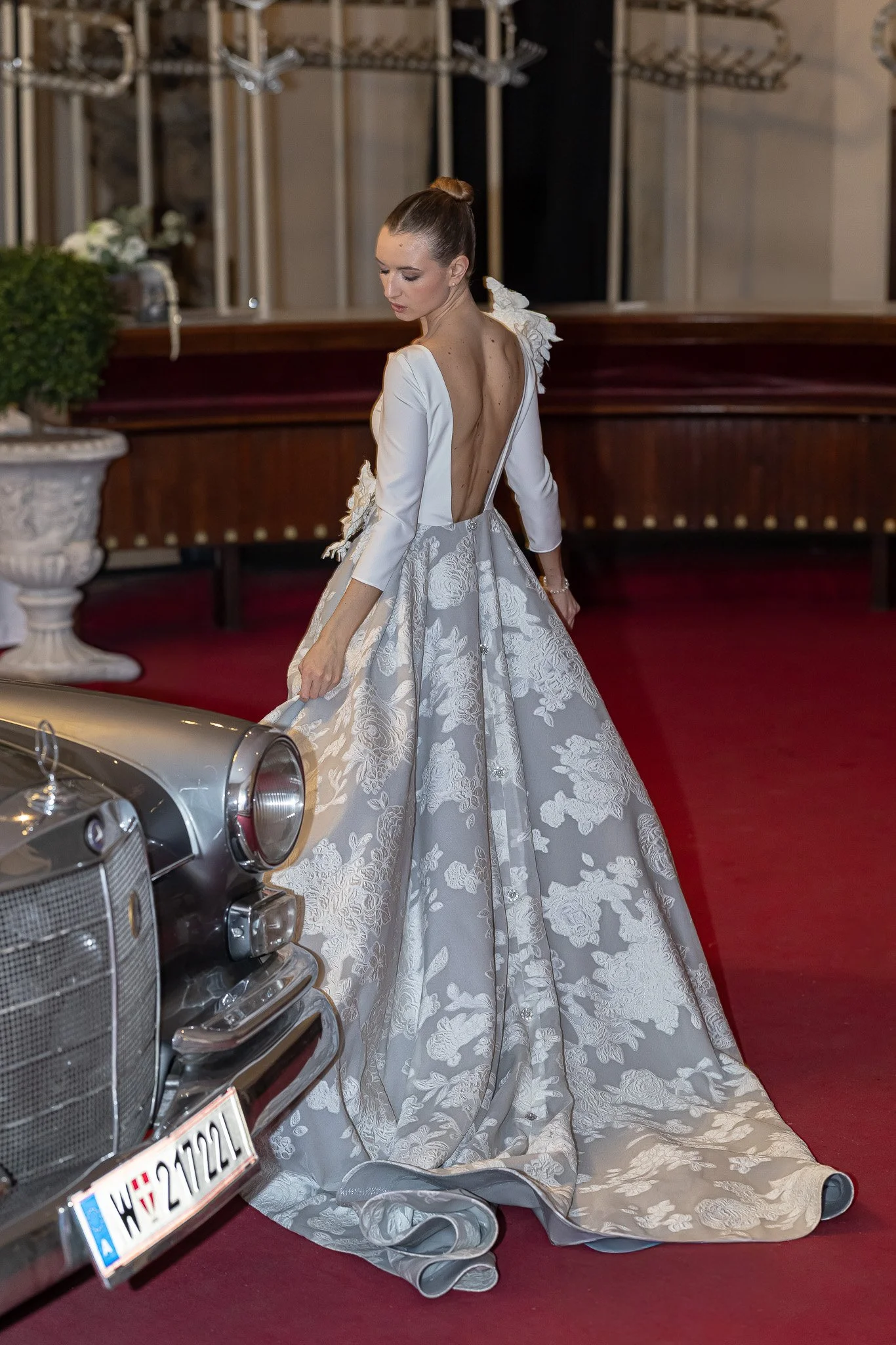 A bride in an elegant white and grey floral wedding gown stands on a red carpet, next to a vintage silver car in Vienna Palais Auersperg, inside a decorated venue with wooden and floral decor.