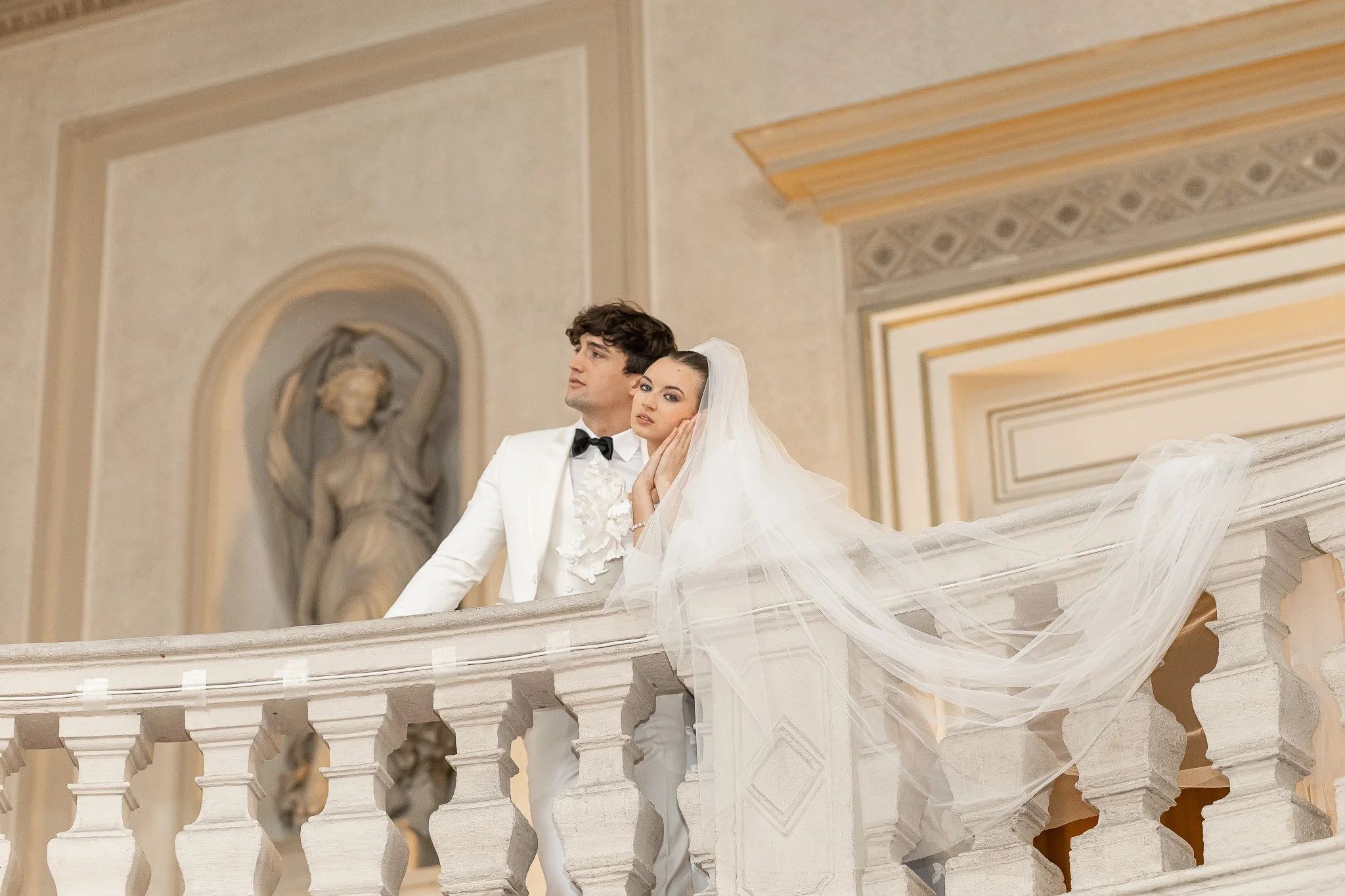 A bride and groom standing together on a balcony inside a grand, ornate building. The bride is wearing a white wedding gown and veil, while the groom is dressed in a white tuxedo with a black bowtie. They are posing together with a statue visible in 