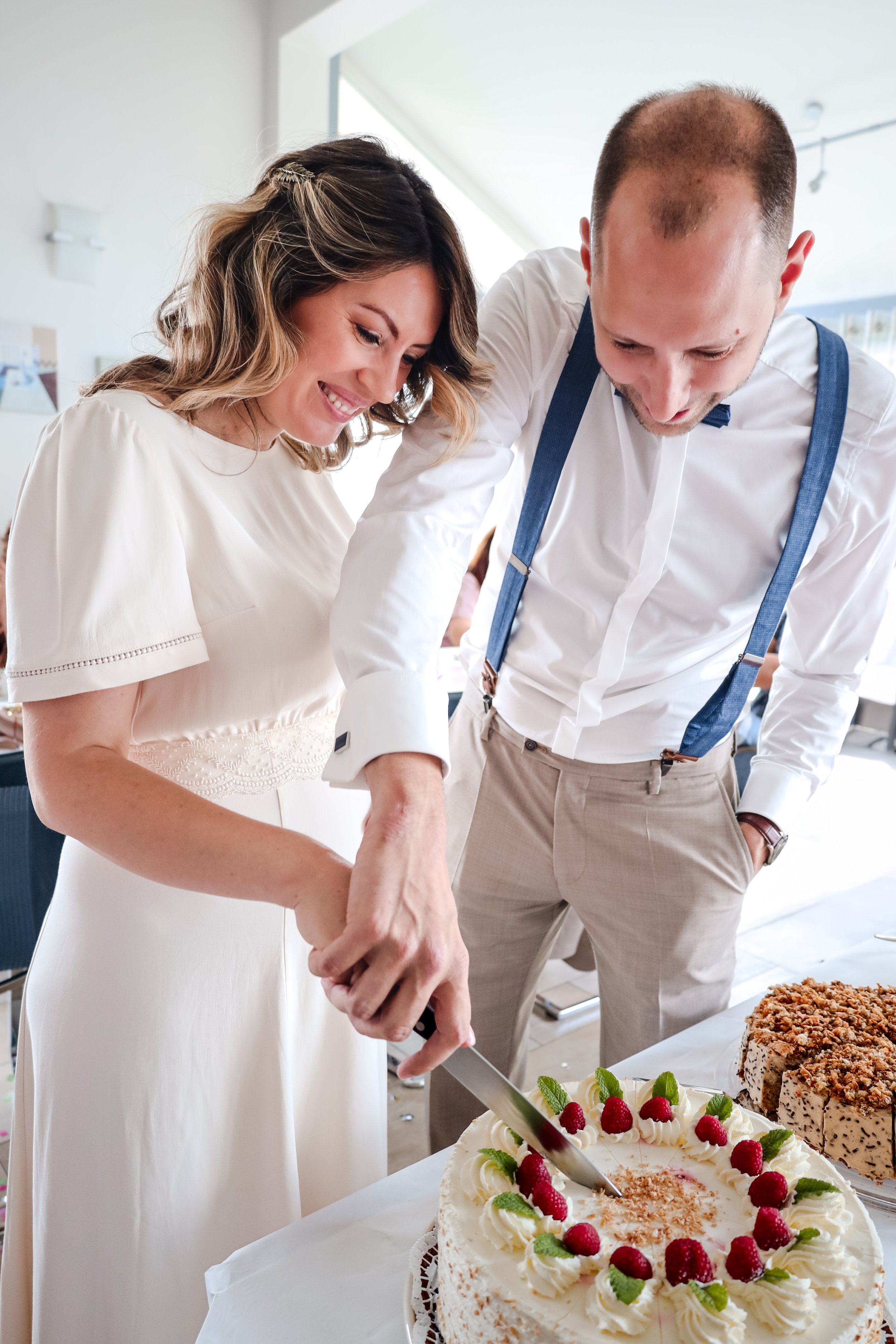 A woman and a man are cutting a wedding cake together, smiling. The woman is wearing a white dress, and the man is dressed in a white shirt with suspenders. The cake is decorated with raspberries, mint leaves, and white frosting.