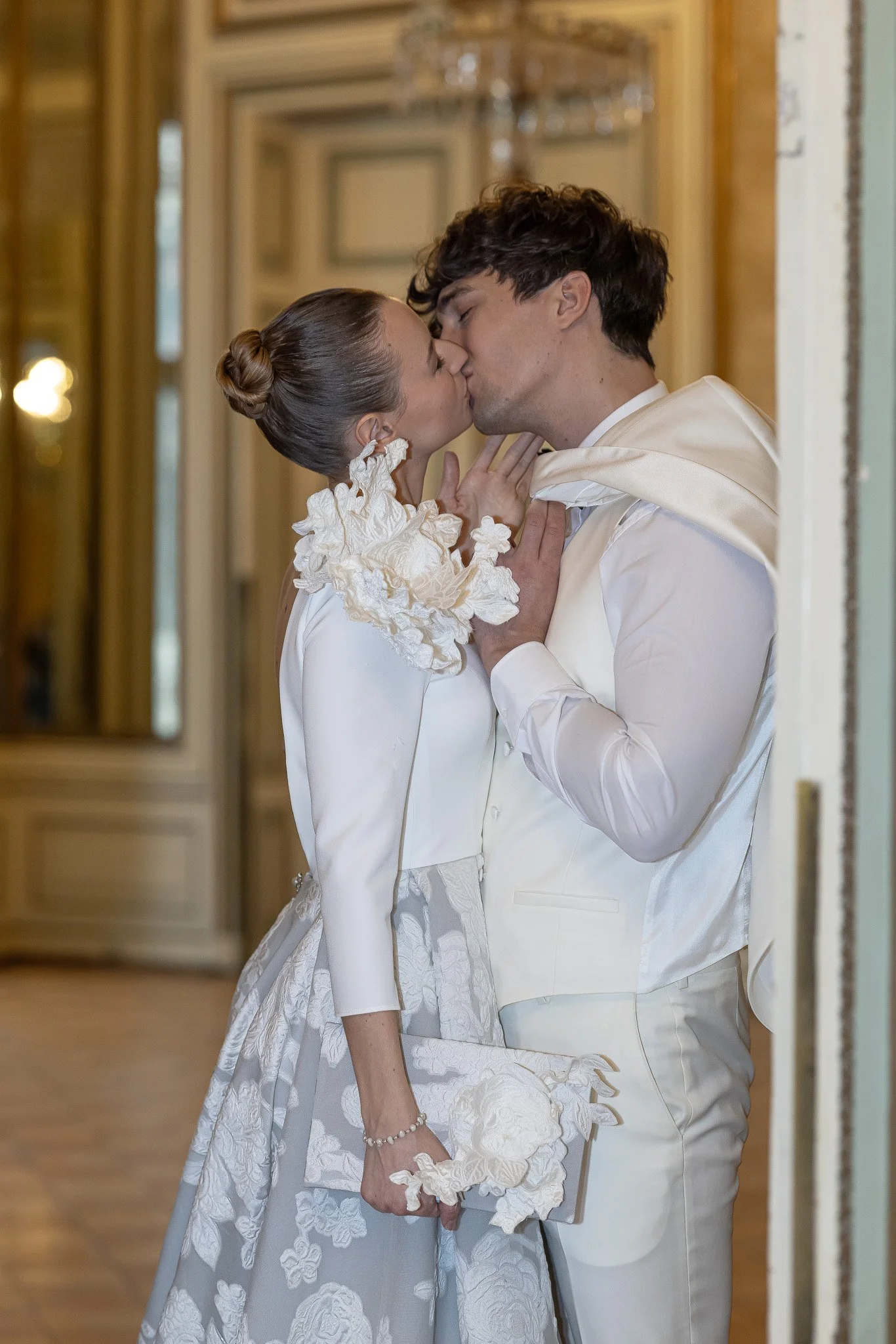 A bride and groom share a kiss in a wedding venue, holding hands and dressed in elegant white wedding attire.