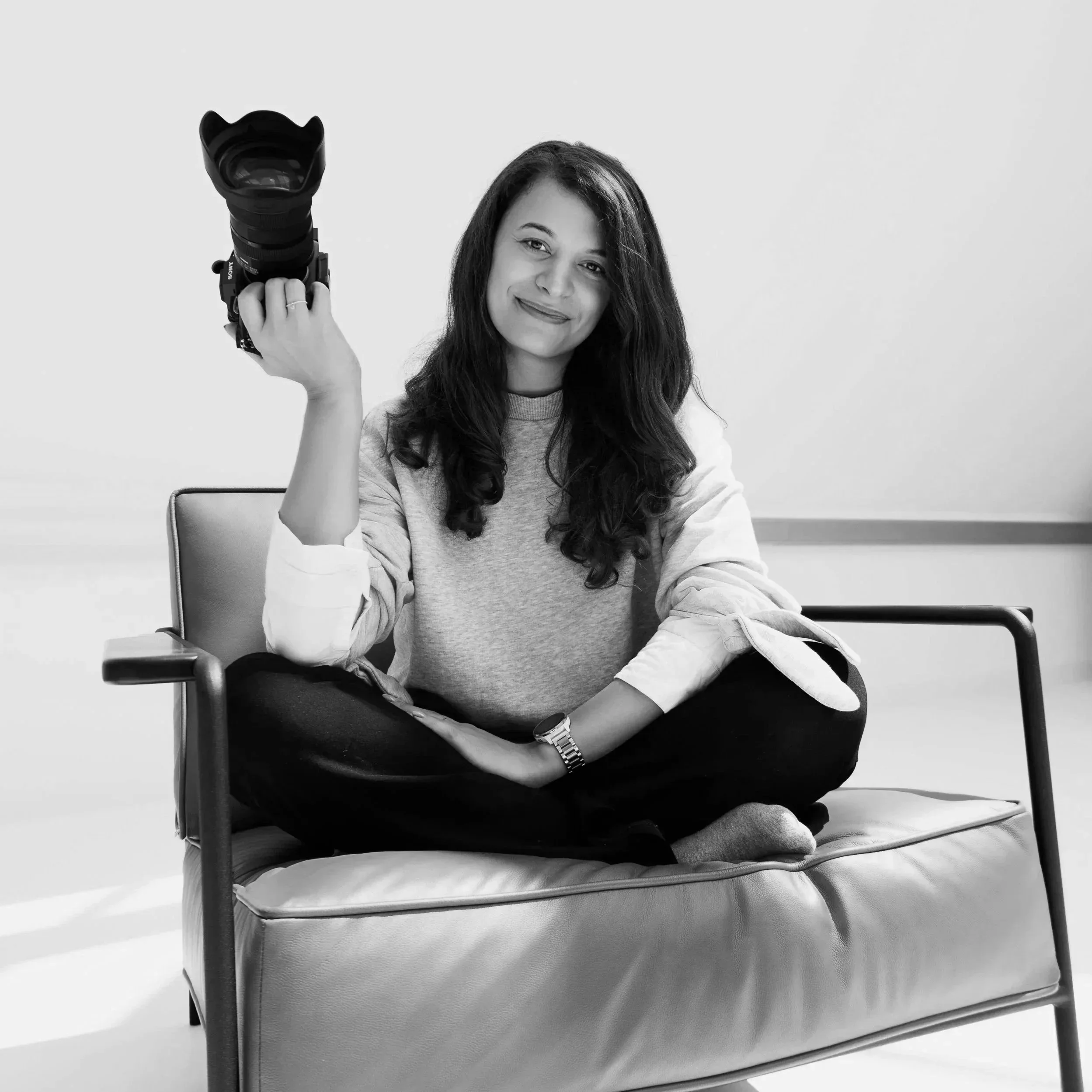 A woman with long hair sitting cross-legged on a couch, holding a camera in her hand, smiling in a black and white photo.