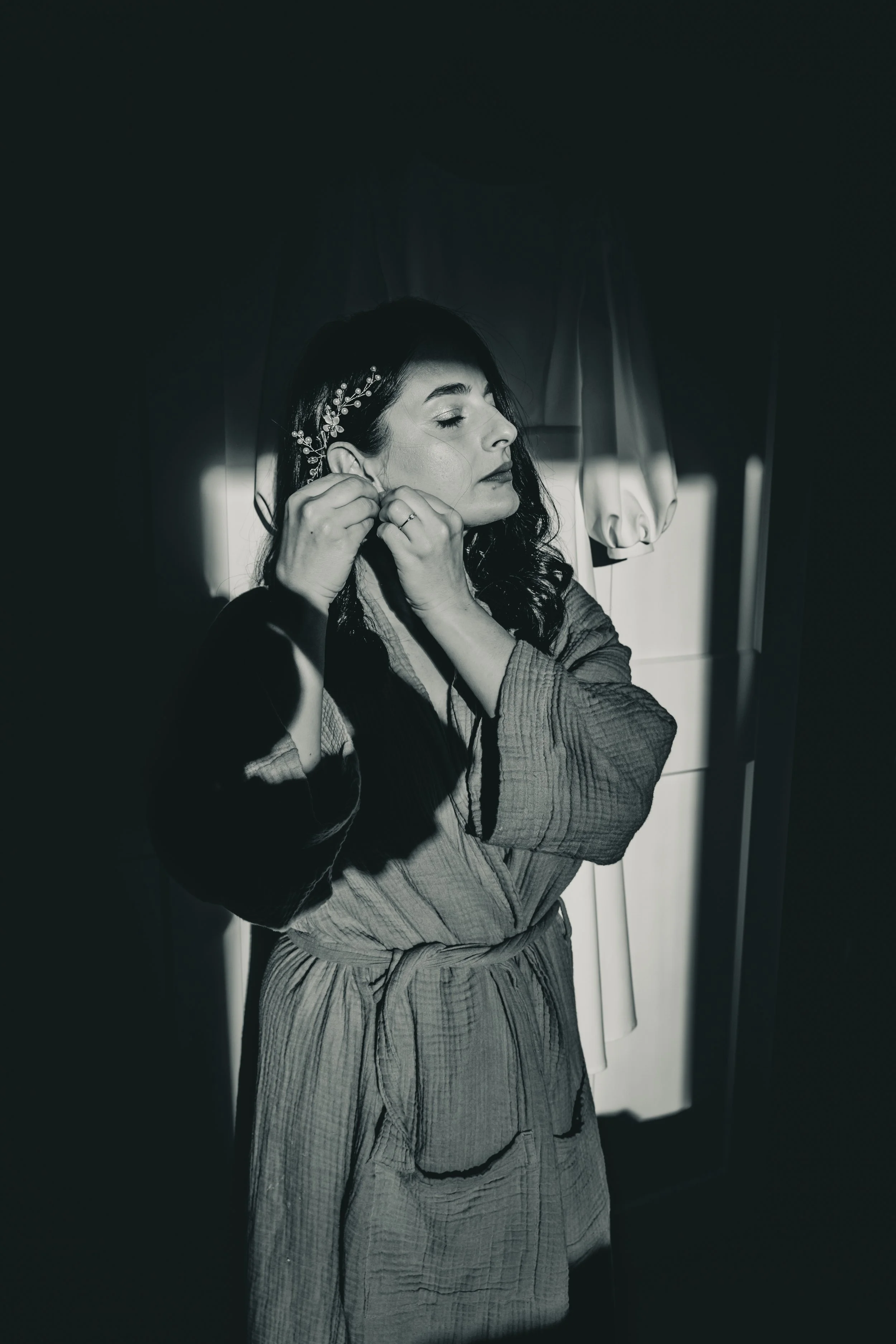 A woman in a bathrobe putting on earrings in a dimly lit room.