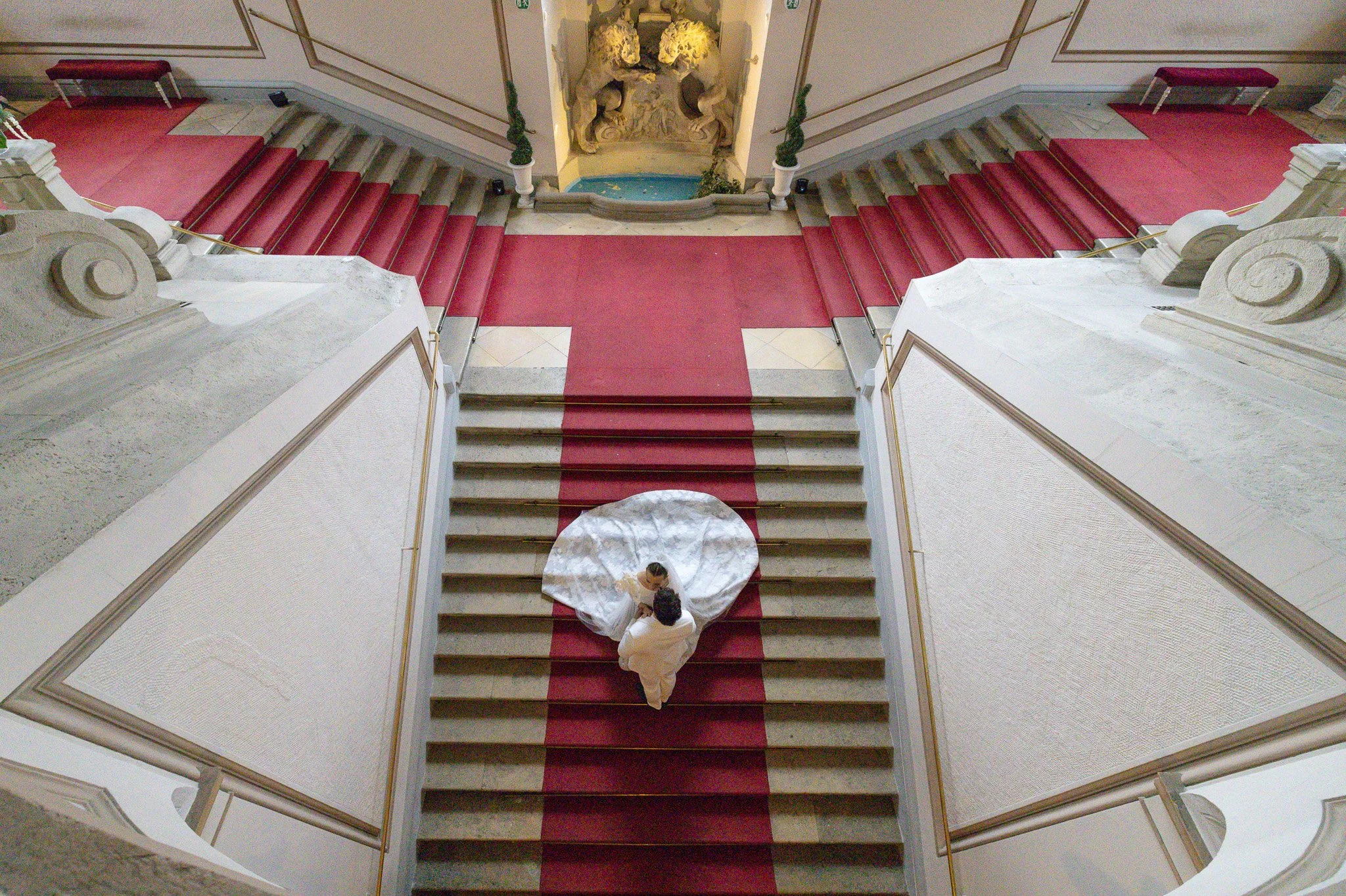 Wedding Photography in Palais Auersperg Vienna. An ornate staircase with red carpet runner, white marble steps, and decorative railings. Two people are standing on the stairs, one holding a white umbrella.