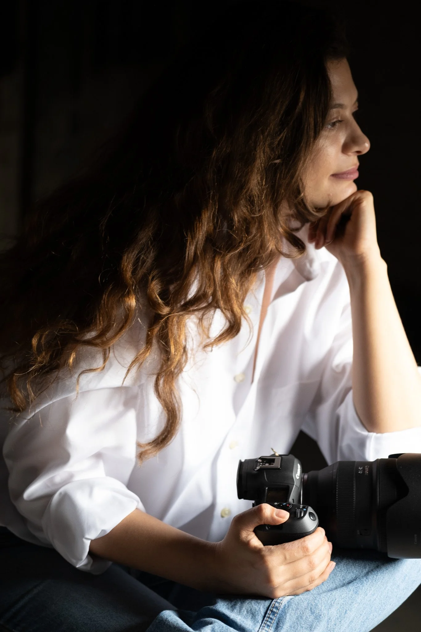 A woman with long wavy brown hair wearing a white shirt, sitting in profile, holding a camera in her right hand, and resting her chin on her left hand.