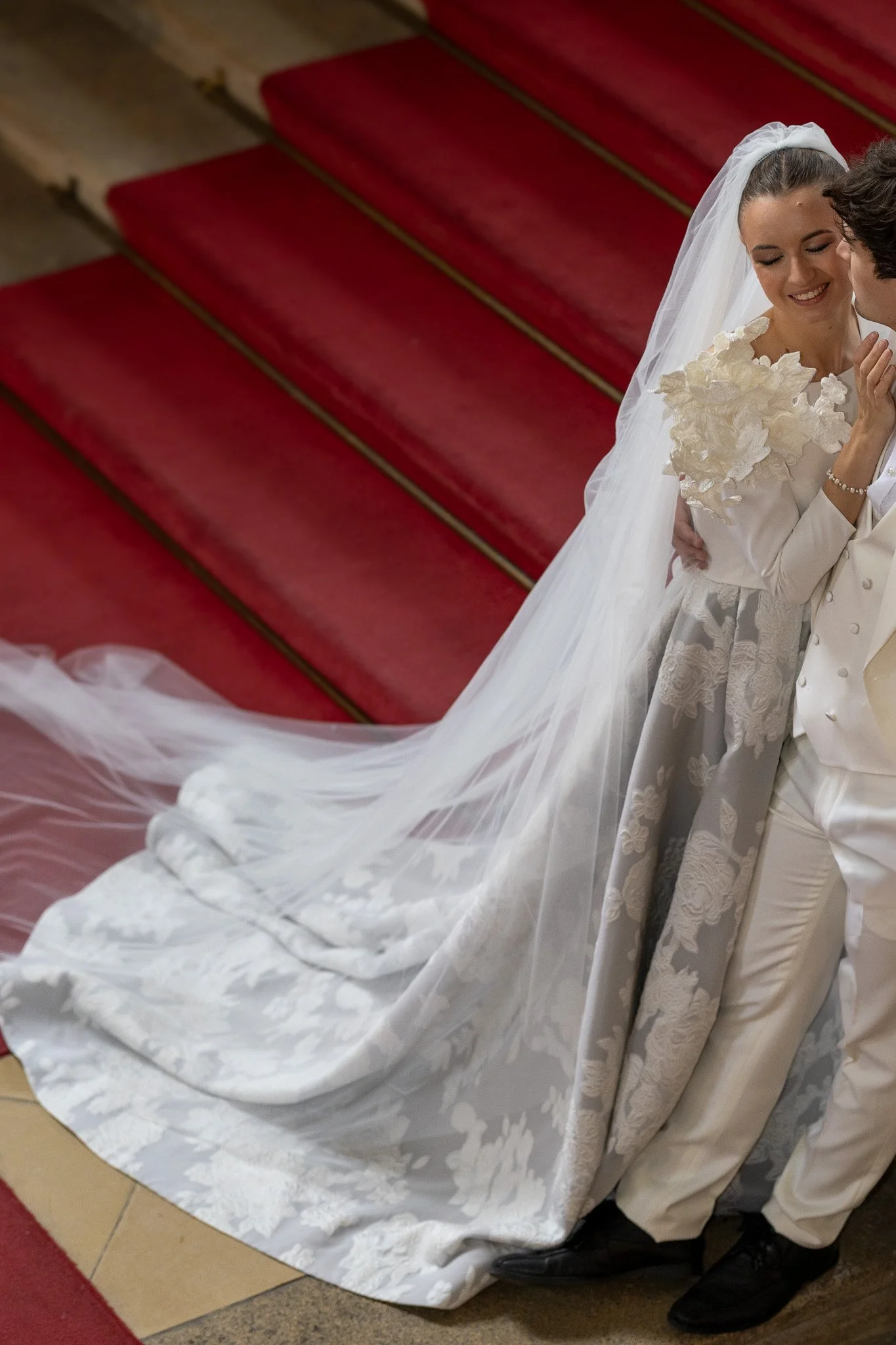 A bride and groom holding each other and smiling during a wedding ceremony, standing on a red carpeted staircase with a white train and veil.