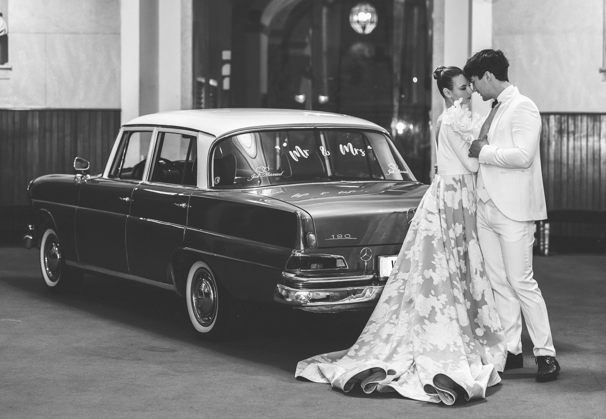 A newlywed couple, dressed in wedding attire, bow their foreheads together in front of a vintage Mercedes-Benz car with wedding decorations on the windshield, inside a building with high ceilings and wooden wall paneling. Vienna, Austria