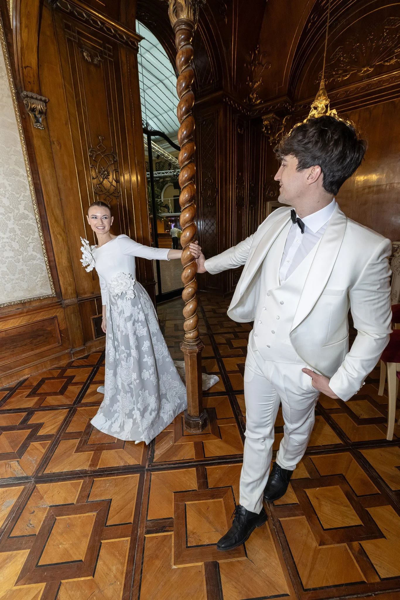 A couple in formal attire, the woman in a white and floral gown and the man in a white tuxedo, dancing or posing in an elegant, wood-paneled room with patterned flooring.