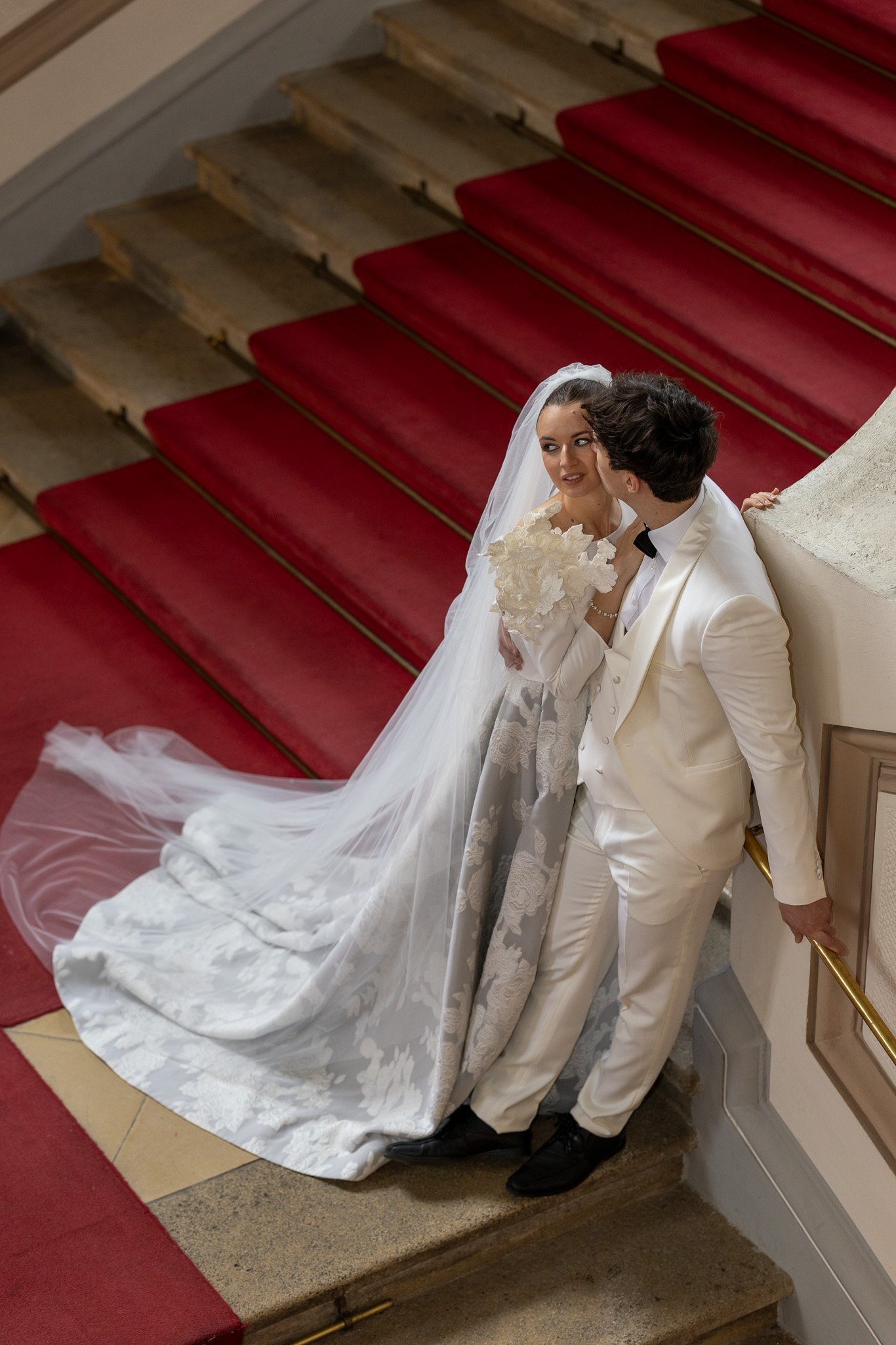 A bride and groom standing close together on a staircase with a red carpet, looking at each other lovingly. Just got married in a palais in Vienna