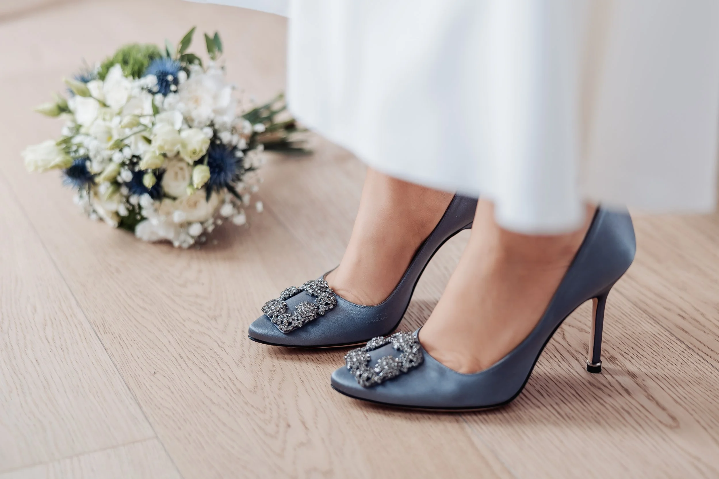 A close-up of a bride wearing gray high heels with decorative buckles and a white dress. A bridal bouquet is on the wooden floor nearby.