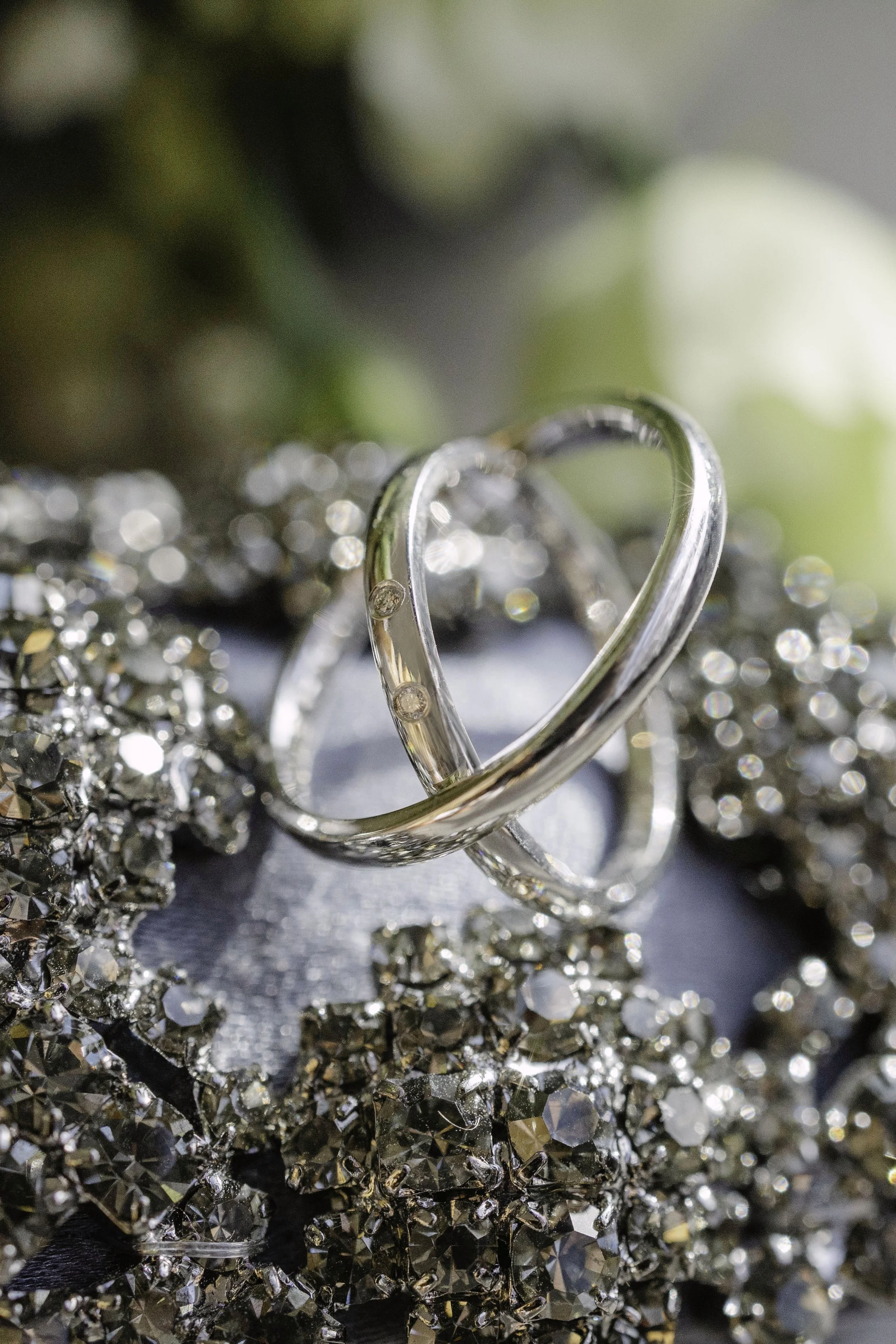 A close-up of intertwined silver rings resting on a bed of glittering, silver-colored rhinestones with a blurred background.