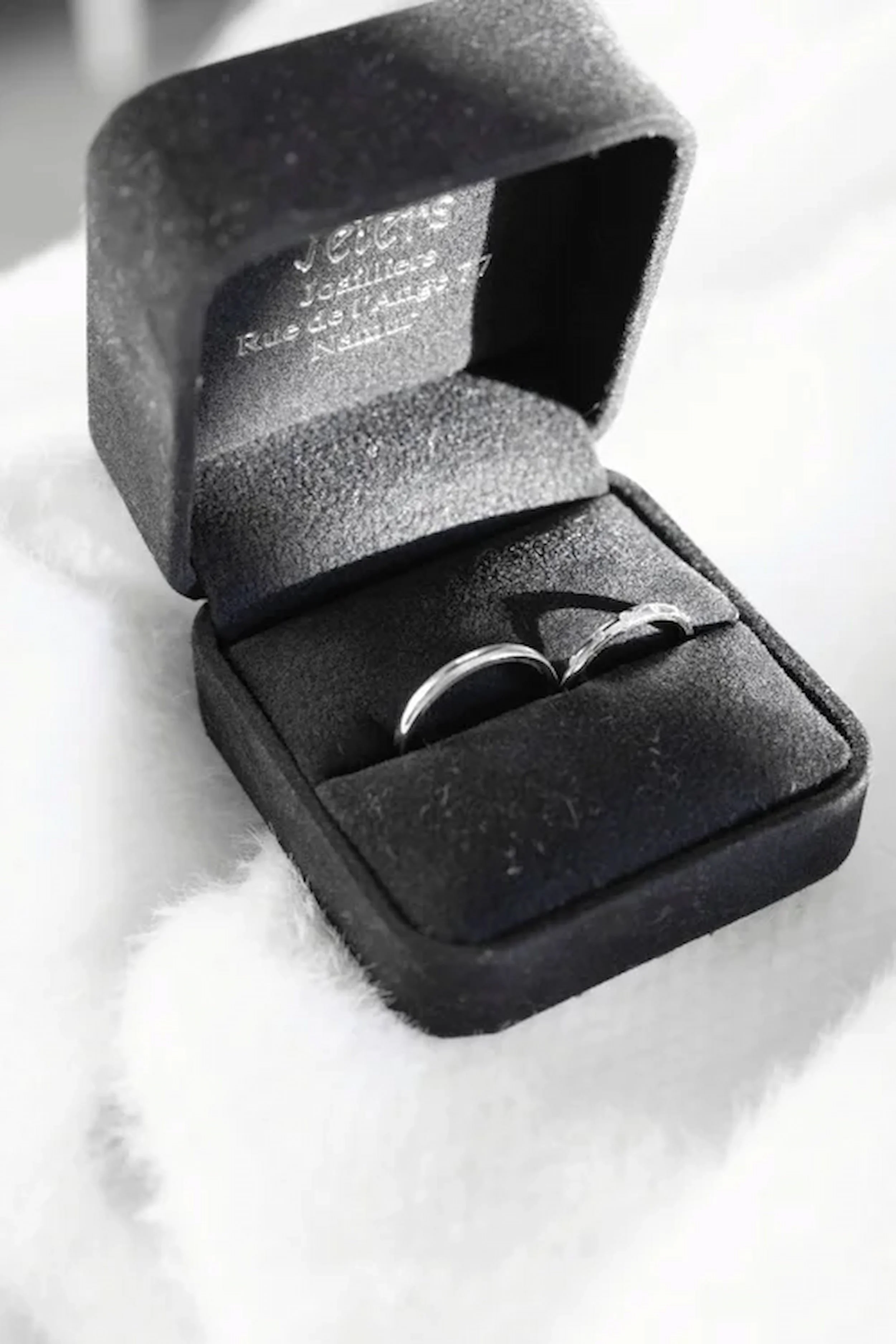 A pair of silver rings in a black ring box lined with black foam, placed on a white, fluffy surface.
