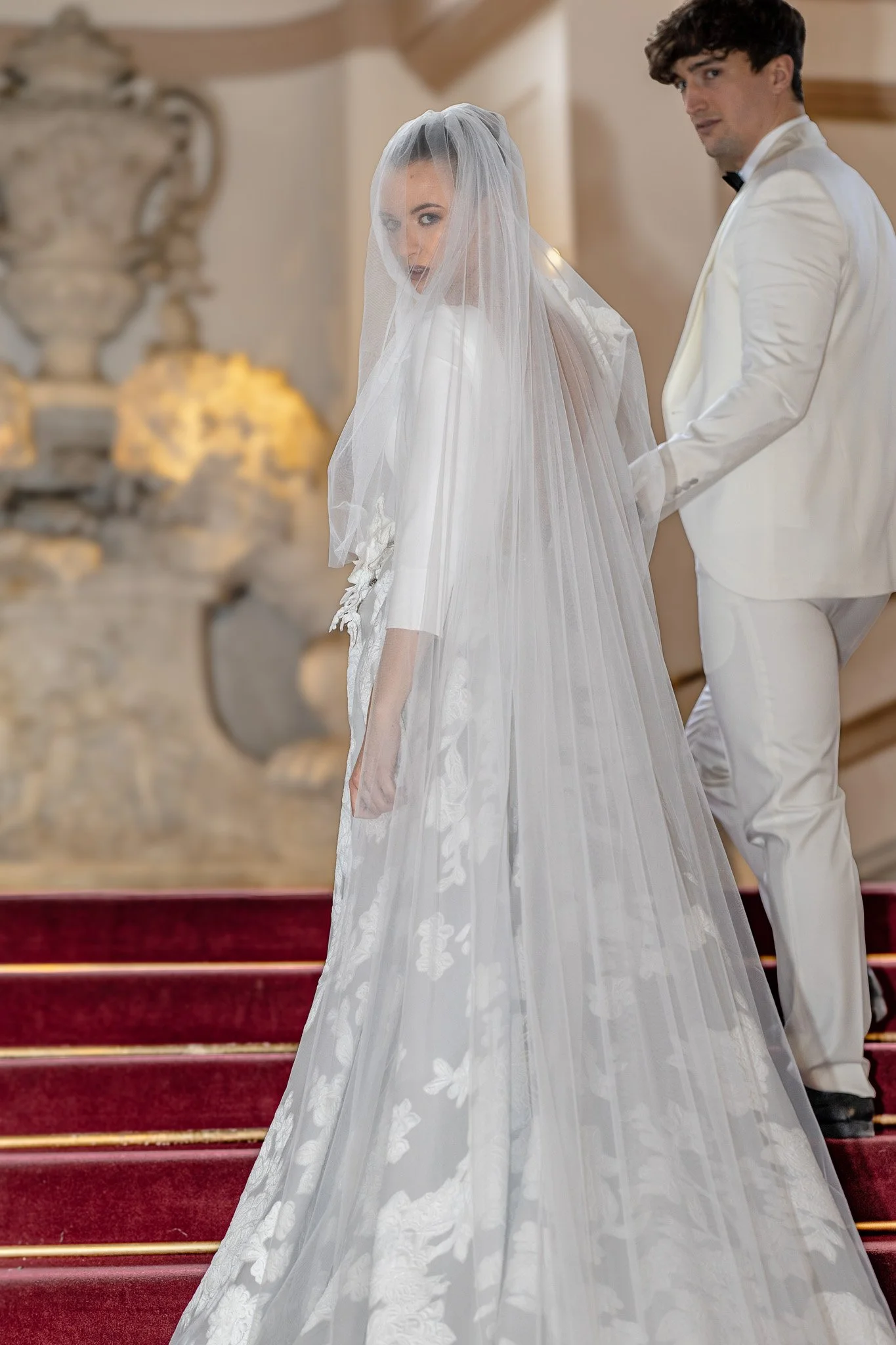A bride in a white wedding dress with a veil standing on red carpet stairs, holding hands with a groom in a white tuxedo. The groom is looking back over his shoulder as they prepare for their wedding ceremony.