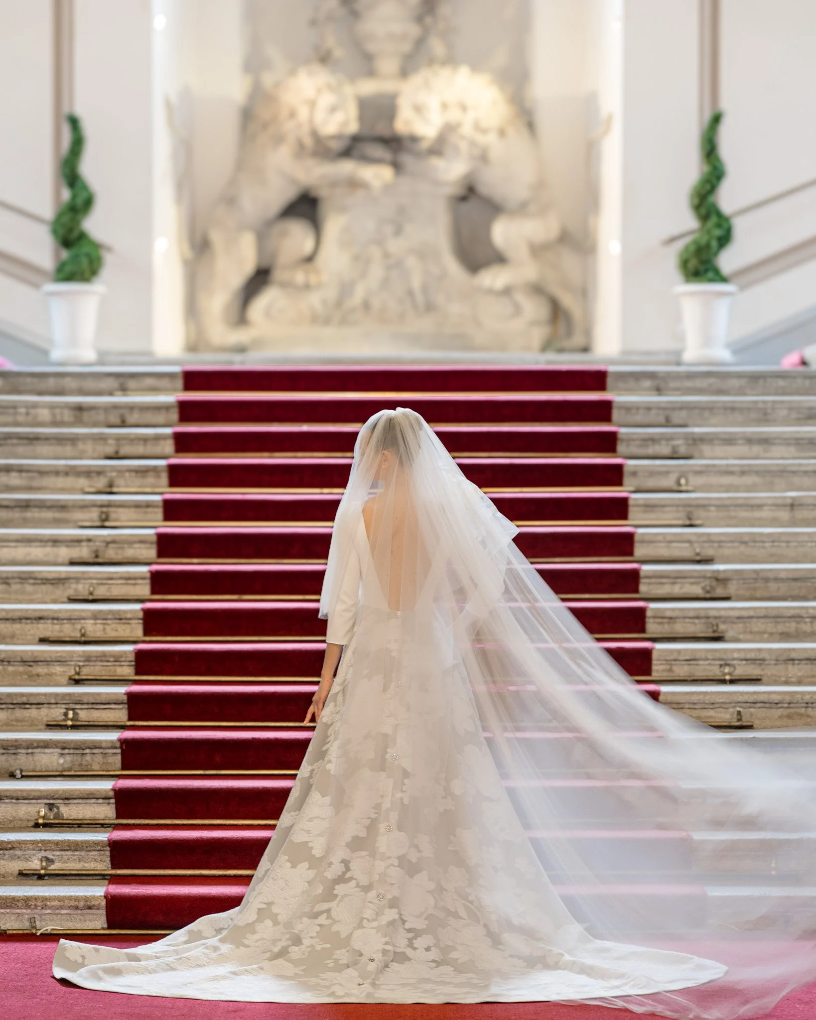 A bride in a white wedding gown with a long train stands at the bottom of a staircase with red carpet runners, facing an ornate stone relief sculpture on the wall.
