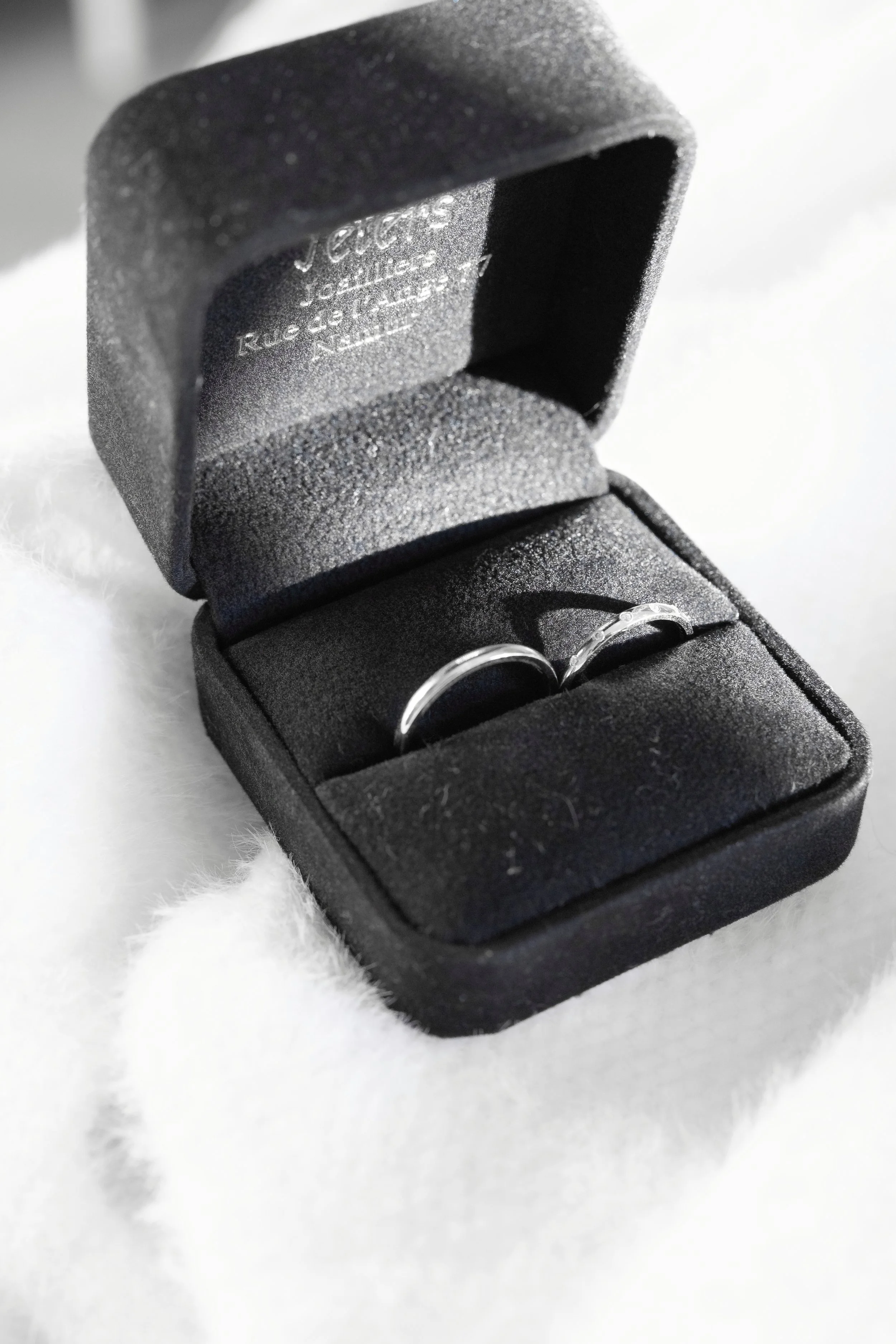 A pair of silver rings in a black ring box lined with black foam, placed on a white, fluffy surface.