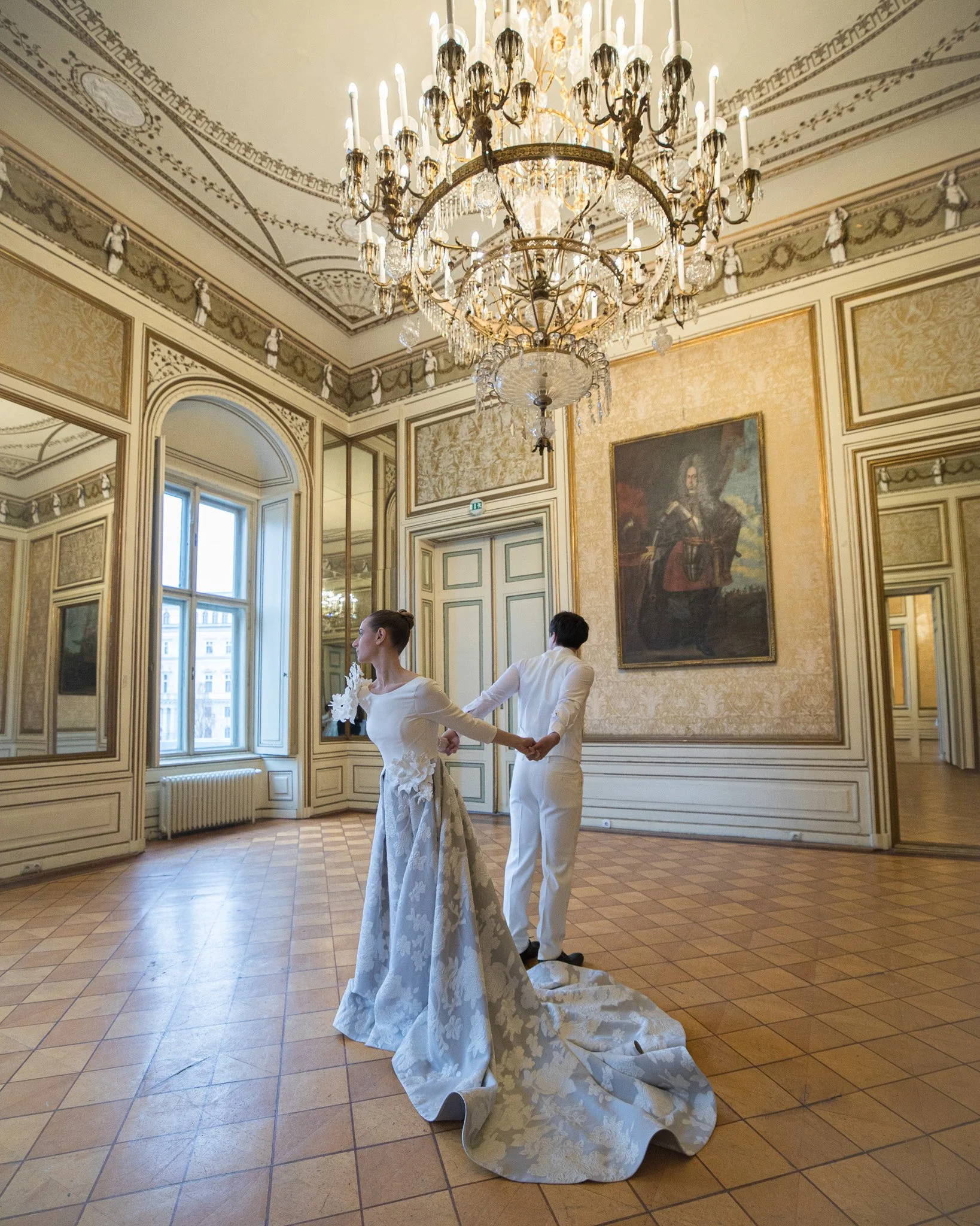 A bride and groom in a historic, ornate room with a large chandelier, gold accents, and a portrait on the wall. The bride is wearing a white gown with a long train, and the groom is dressed in white. They are holding hands behind their backs.